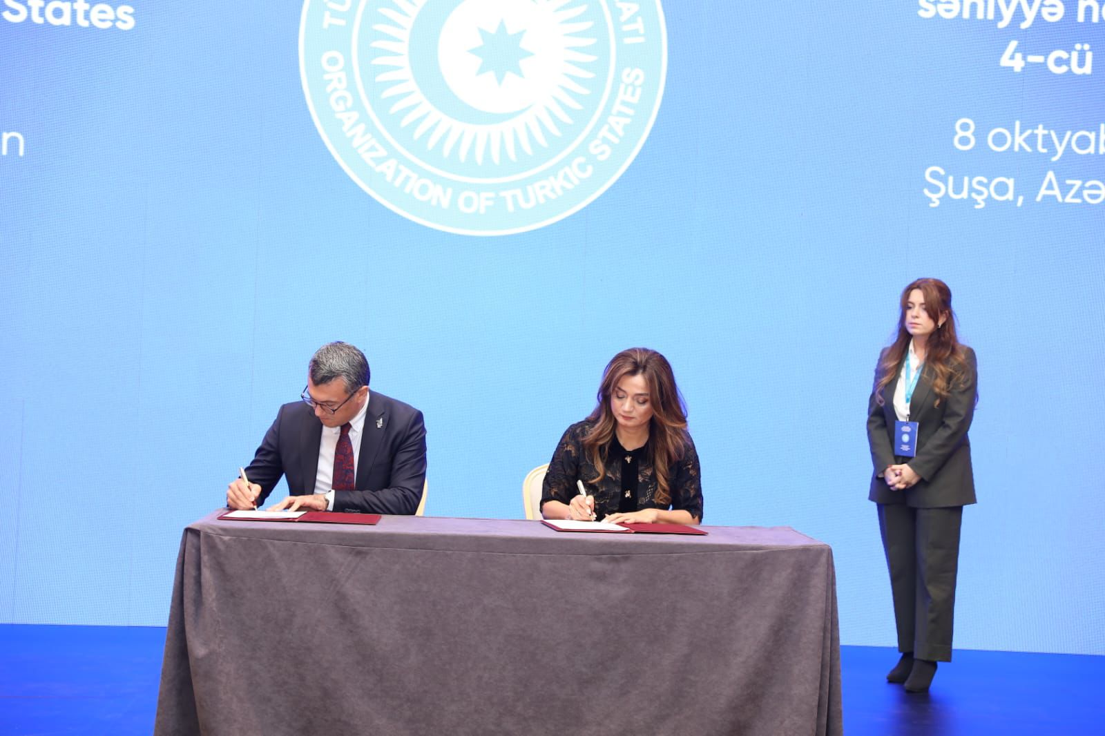 Two individuals are signing documents at a table, with a blue background displaying the logo of the Organization of Turkic States.