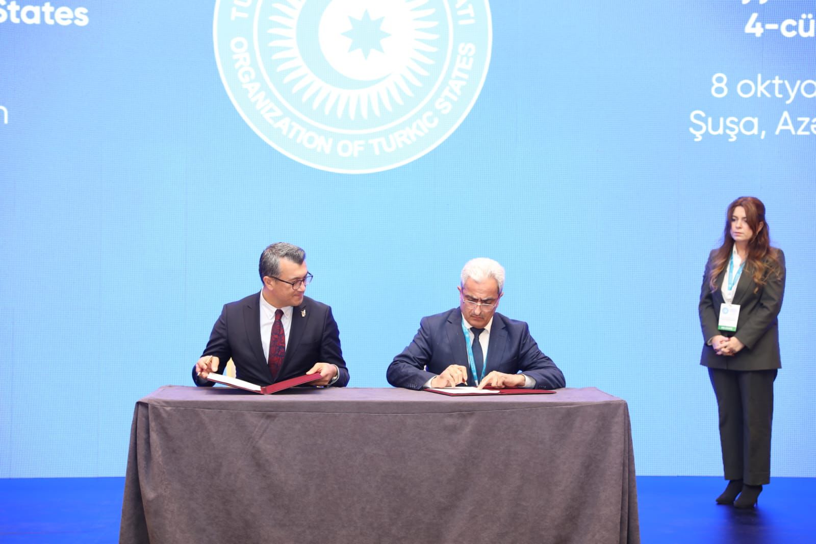 Two officials are seated at a table, signing documents, with a woman standing in the background. A blue backdrop features the logo of the Organization of Turkic States.