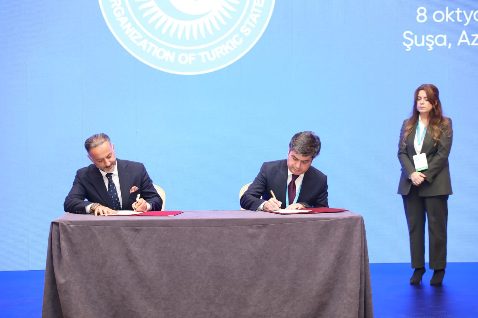 Two men are signing documents at a table, with a woman standing in the background. A blue screen displays the logo of the Organization of Turkic States.