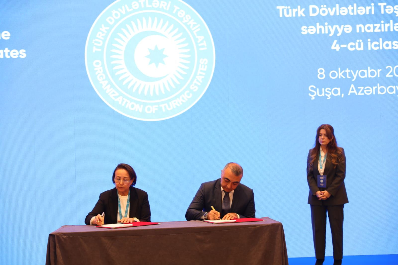 Two officials are signing documents at a conference, with a blue backdrop displaying the logo of the Organization of Turkic States.