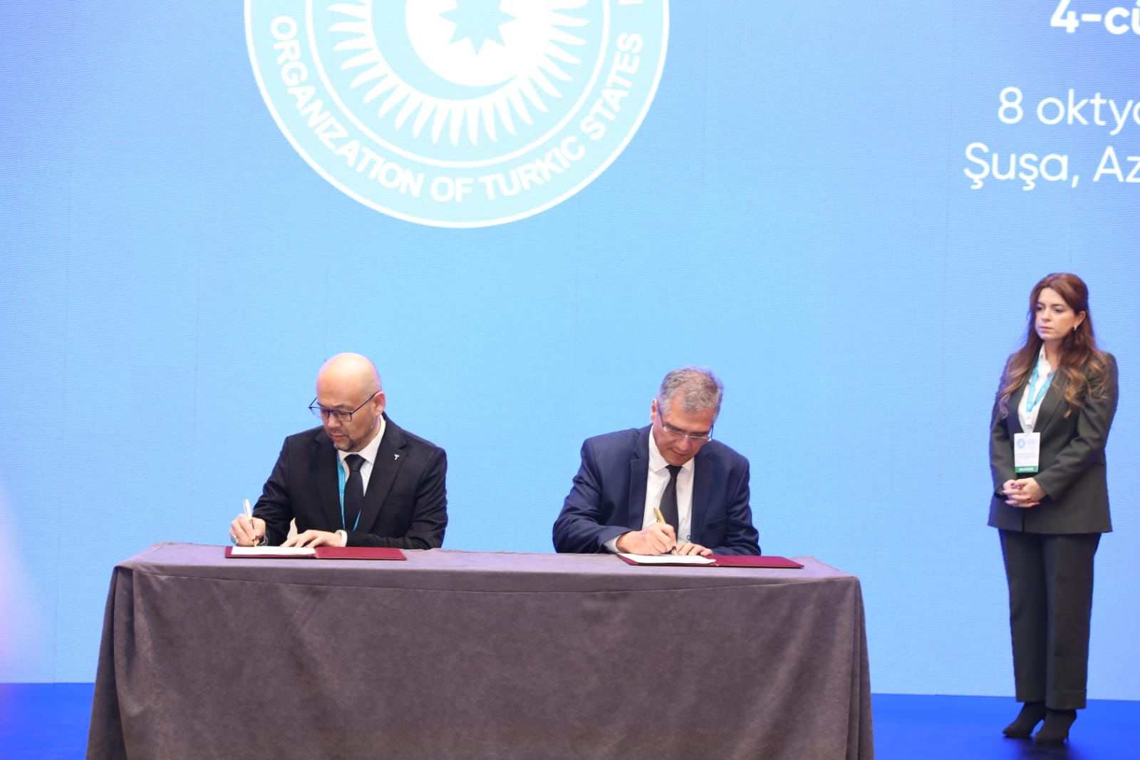 Two men in suits are signing documents at a table, with a woman in formal attire standing nearby. A blue backdrop features the Organization of Turkic States logo.