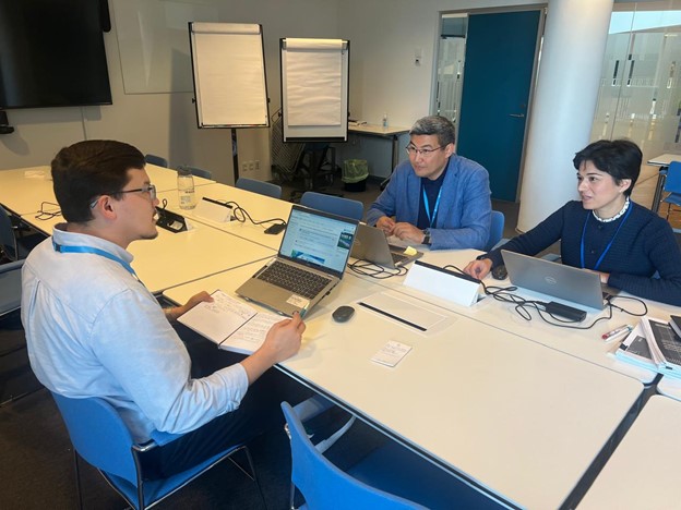 Three people are seated at a table in a meeting room, discussing with laptops and notes in front of them.