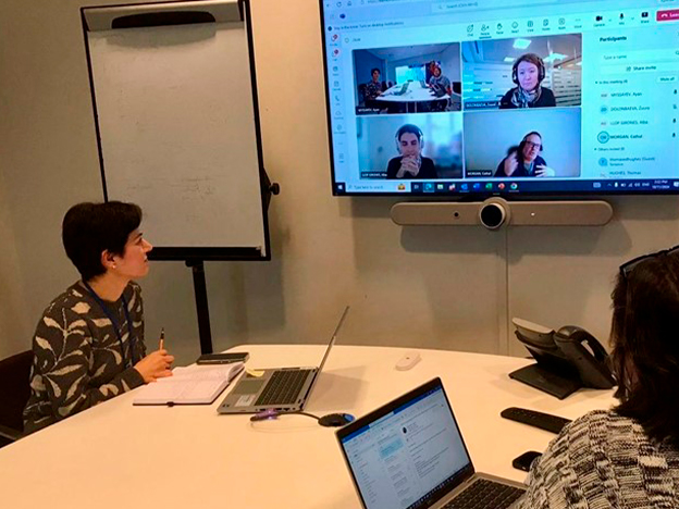 A group meeting in a conference room with a person taking notes, laptops open, and a large screen displaying remote participants.