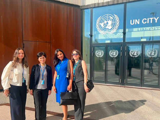 Four women stand outside UN City, smiling and wearing identification badges, with the UN logo visible on the building.