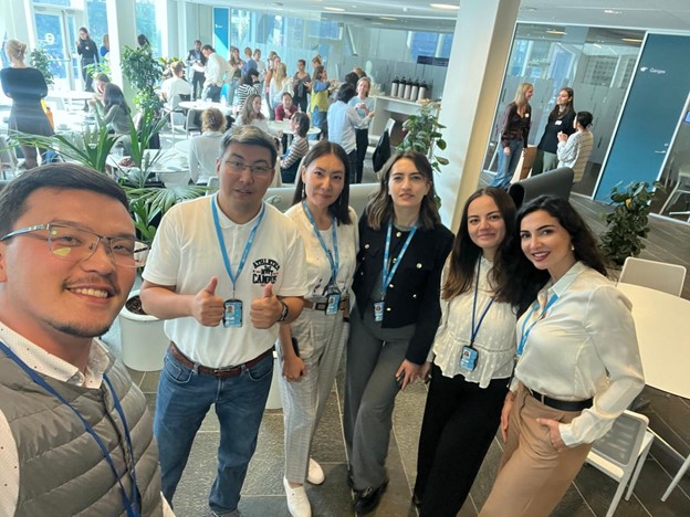 A group of six people poses for a selfie in a modern office space, with plants and colleagues in the background.