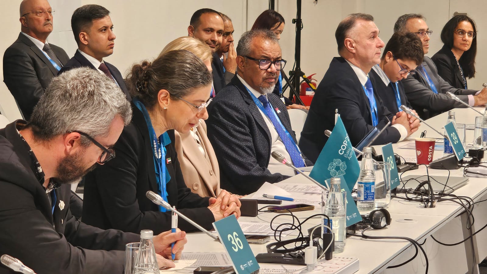 A group of professionals seated at a conference table, engaged in discussion, with flags and nameplates visible.