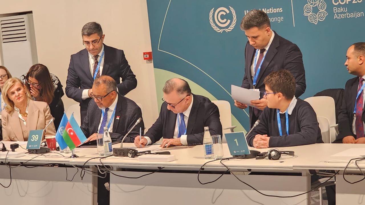 A group of officials at a conference table, with flags and documents, engaged in discussions and signing agreements.