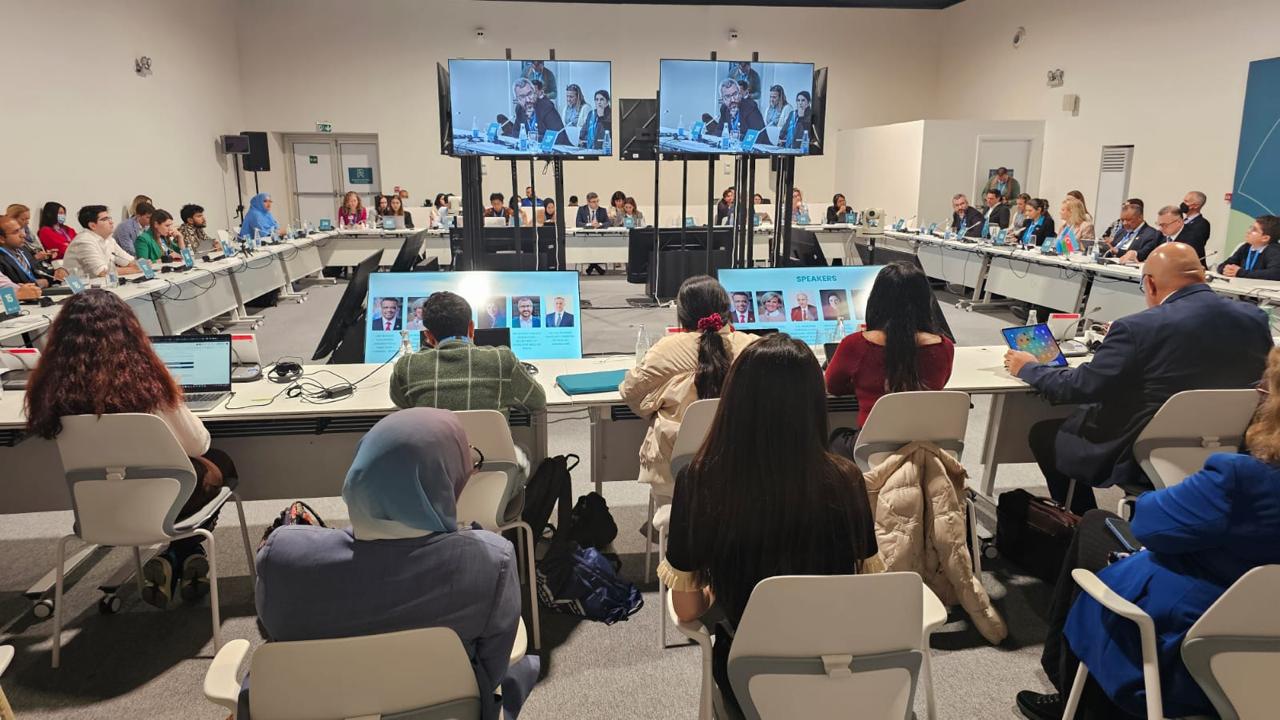 A conference room with participants seated at tables, facing screens displaying speakers. Laptops and notes are visible.