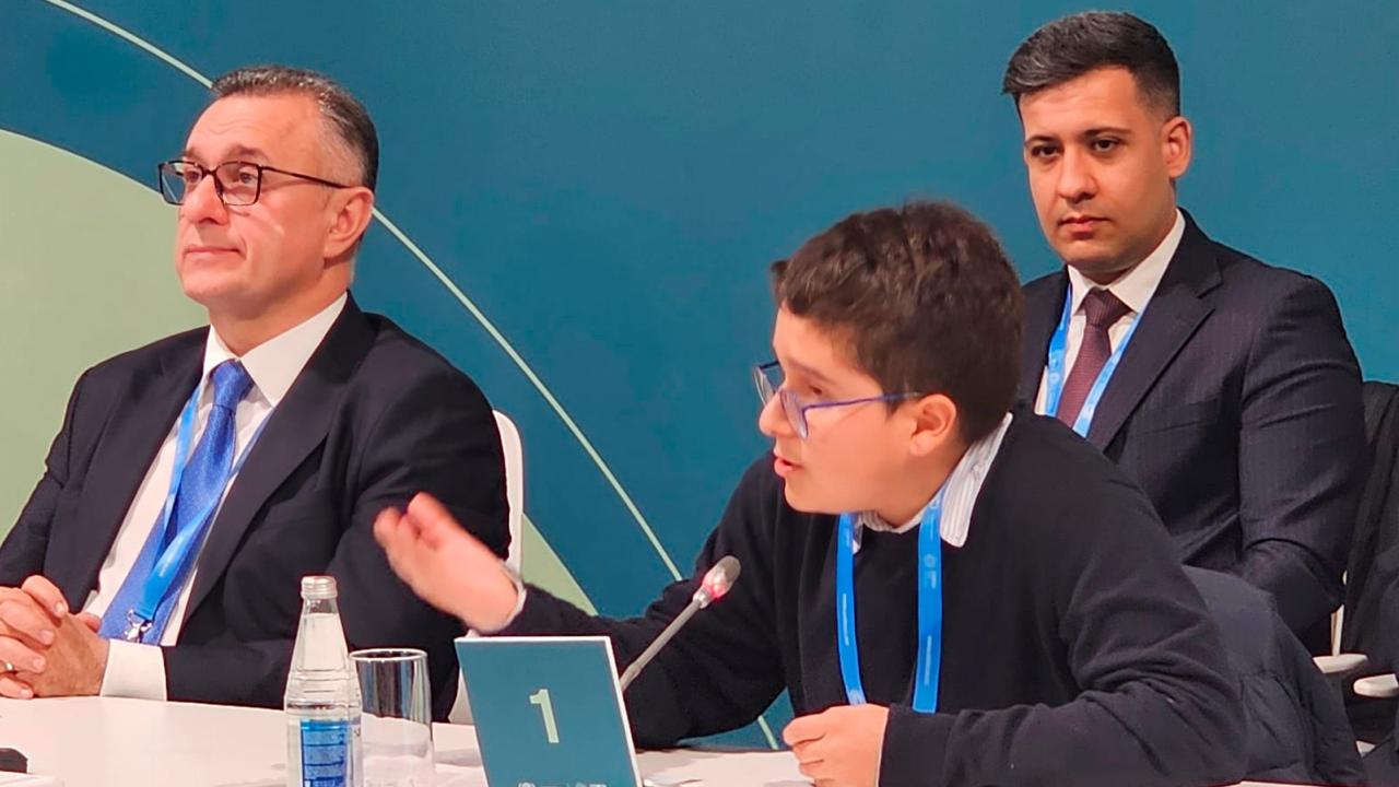 A young boy speaks at a conference table, flanked by two men in formal attire, with a blue backdrop.