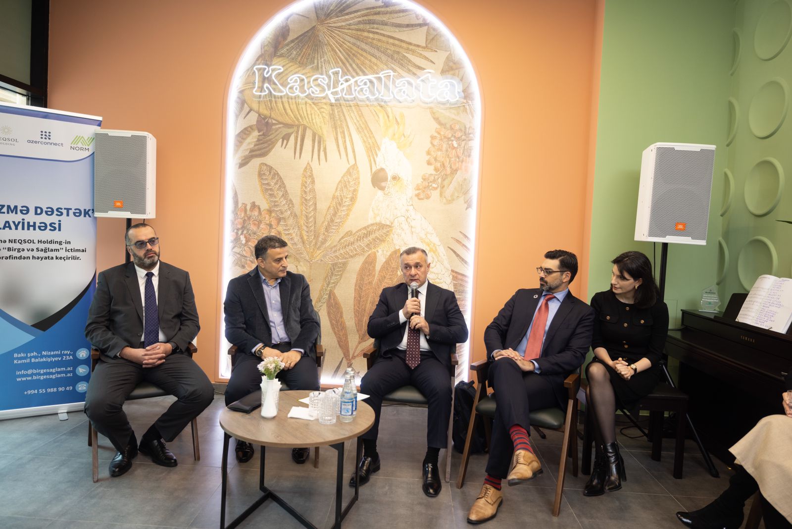 A panel discussion featuring five individuals seated in a modern setting, with a decorative wall and a banner in the background.