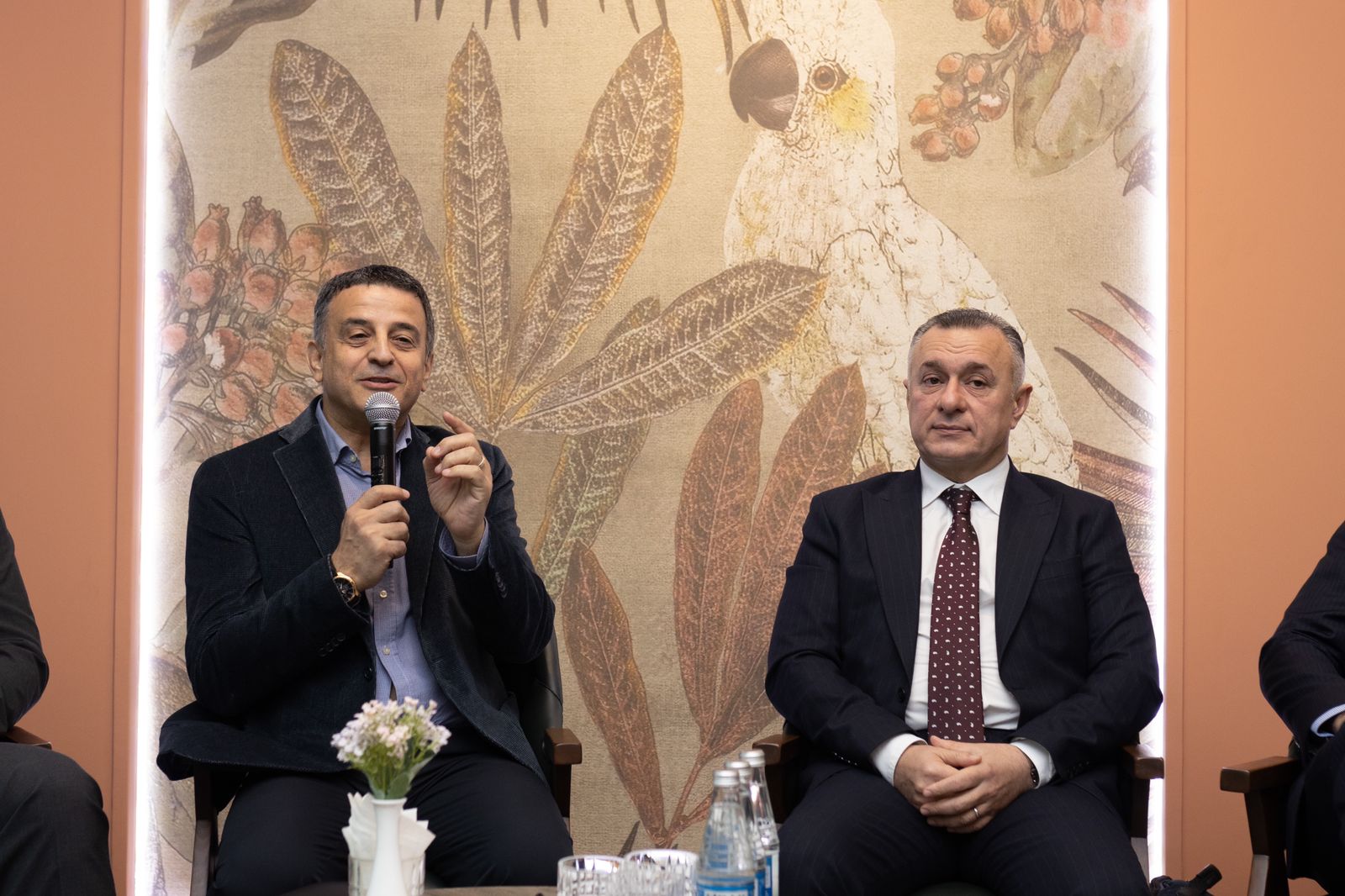 Two men are seated at a table, one speaking into a microphone. A decorative background features leaves and a bird.