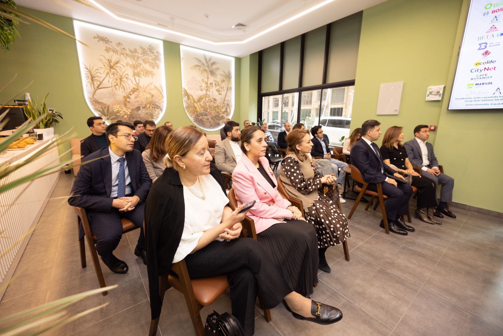 A group of people seated in a modern room with greenery and a large screen, engaged in a presentation or meeting.