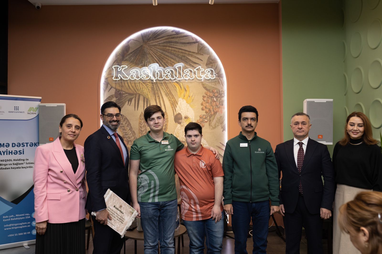 A group of six people stands together in a café, with a decorative wall and a sign reading "Kashalata" in the background.