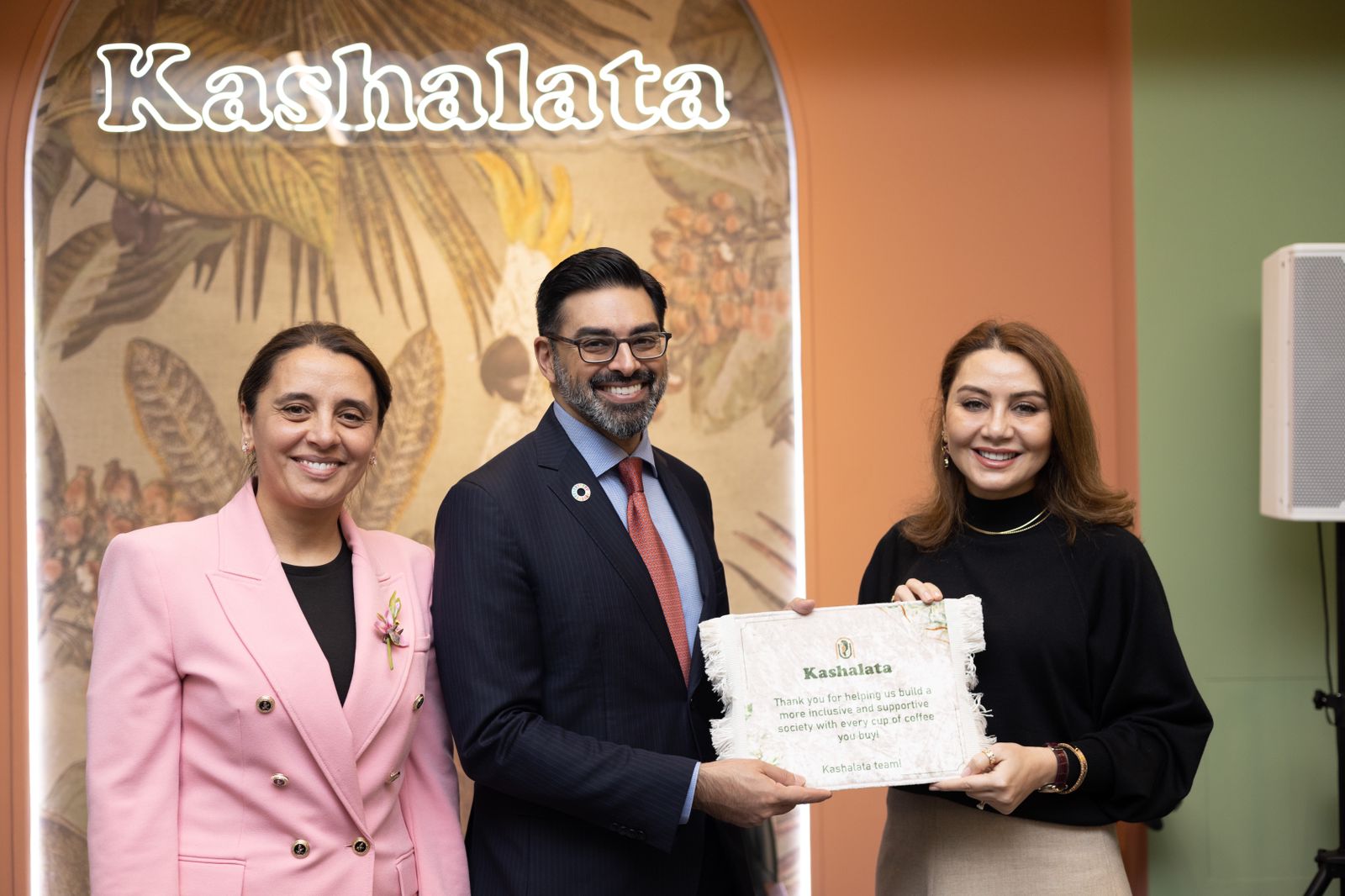 Three individuals pose together, holding a certificate in front of a decorative backdrop with the name "Kashalata."