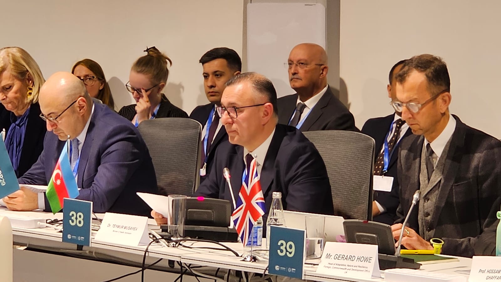 A group of officials seated at a conference table, discussing with flags and nameplates visible.