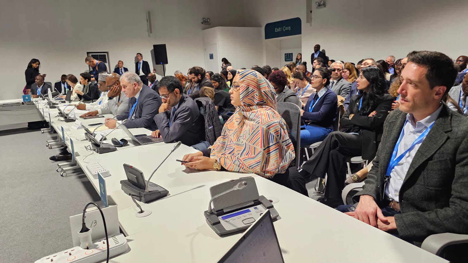 A diverse group of people seated at a conference table, engaged in discussion, with some using laptops and taking notes.