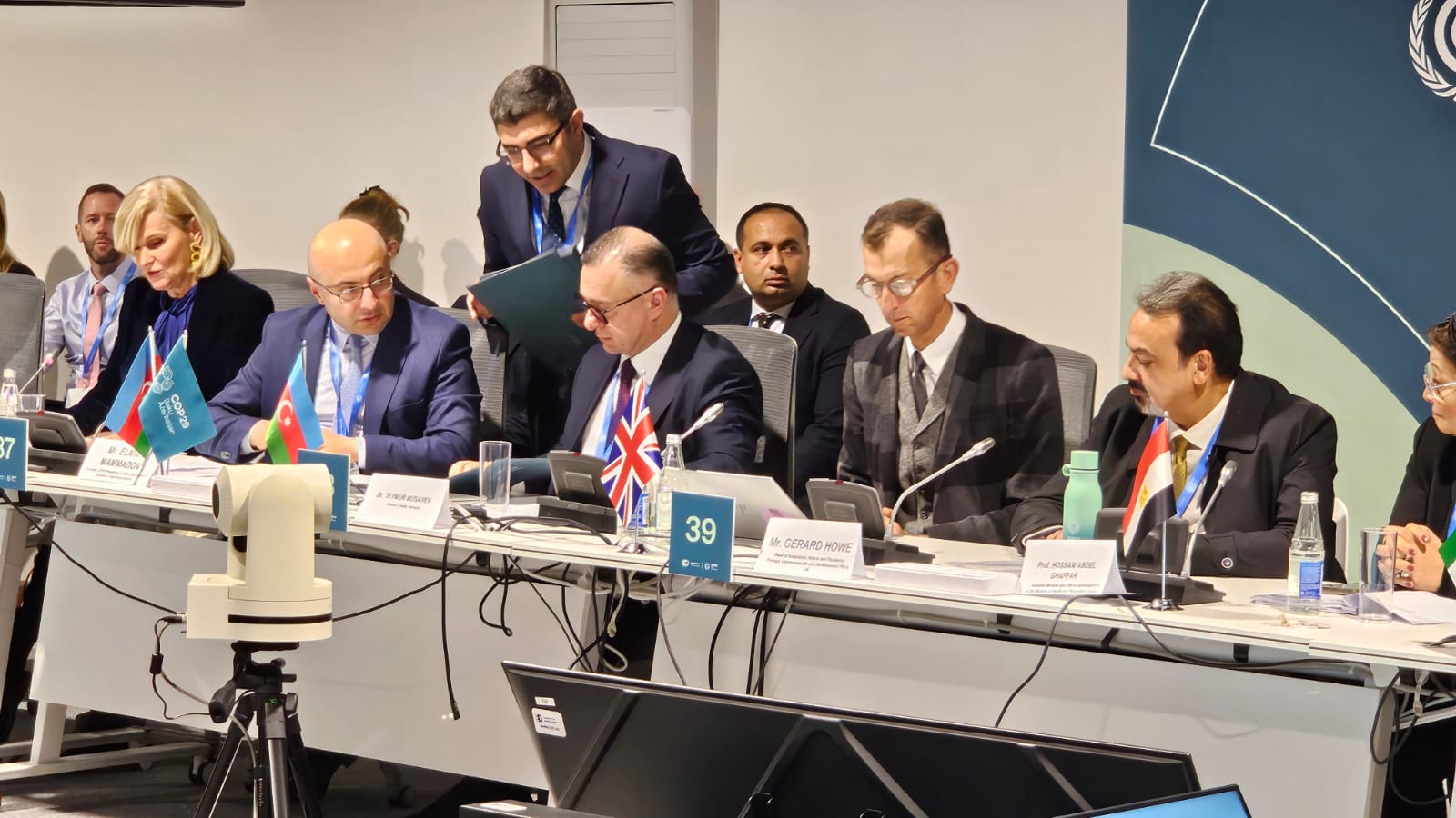 A group of delegates at a conference table, discussing with flags and nameplates visible. Various individuals are engaged in conversation.