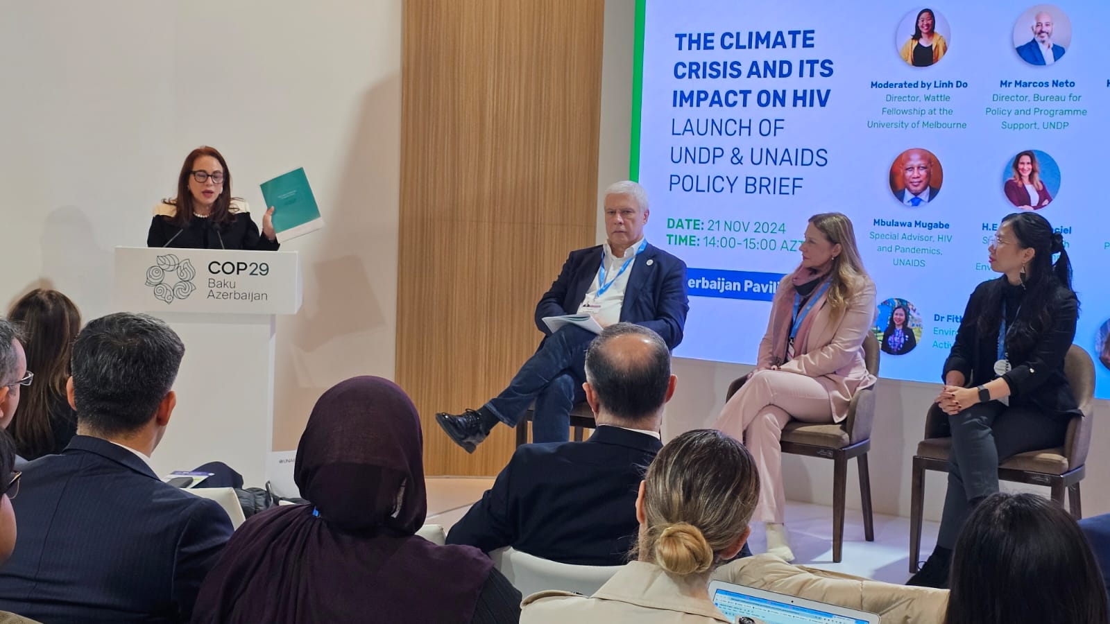 A speaker presents a policy brief at COP29 in Baku, Azerbaijan, with panelists seated nearby. A screen displays event details.