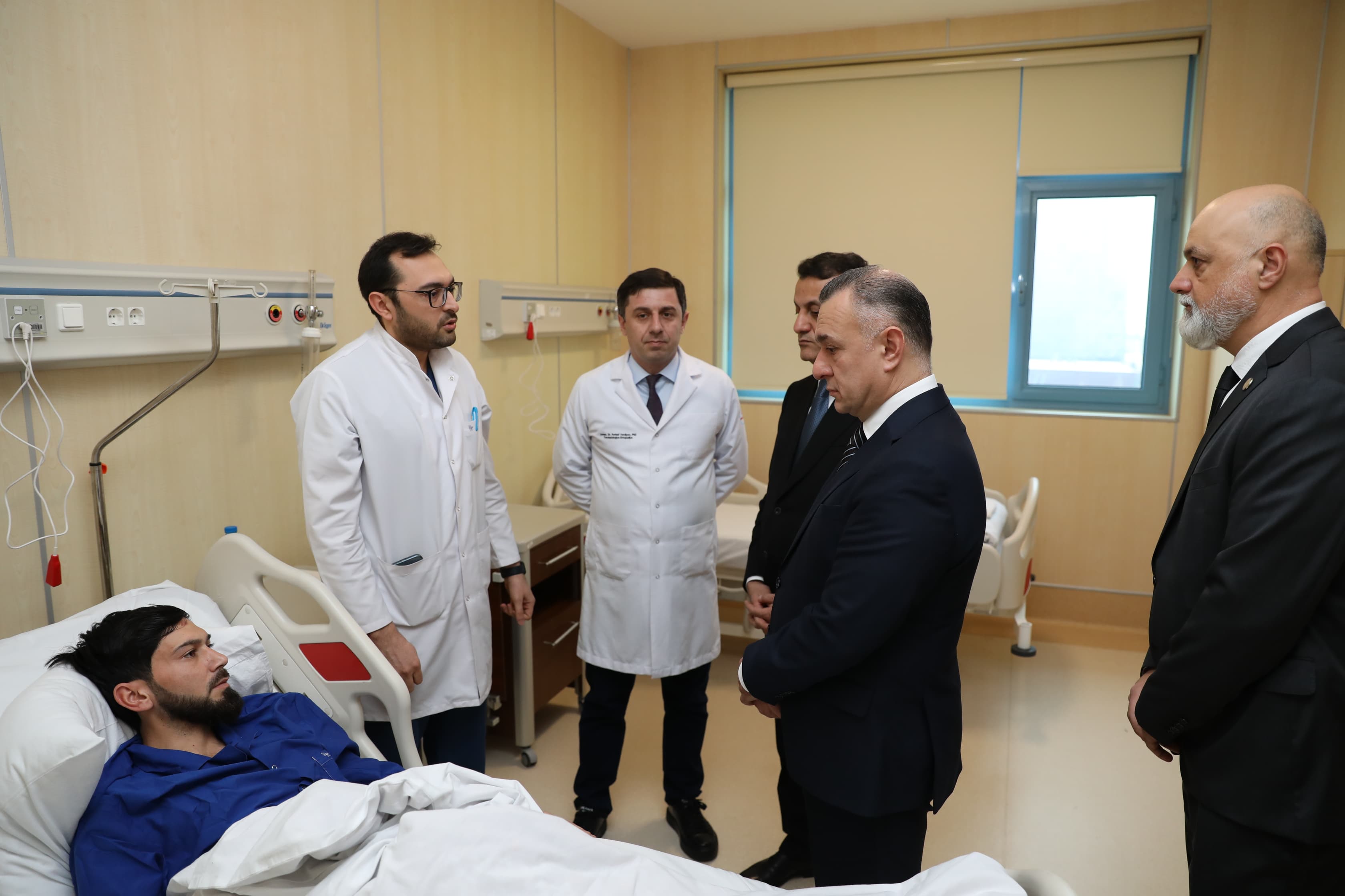 A patient in a hospital bed is visited by several medical professionals and officials, discussing his condition in a clinical setting.