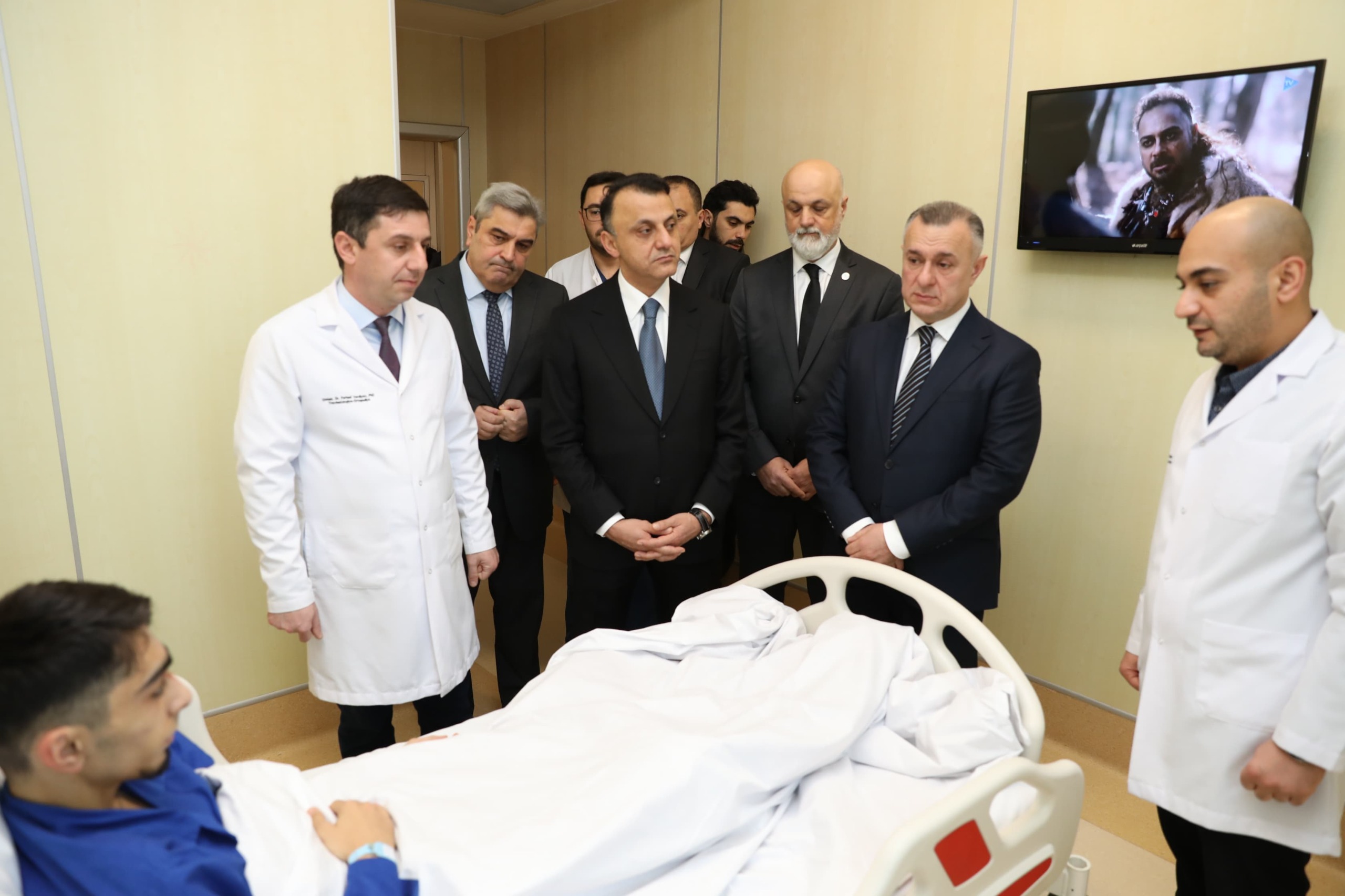 A group of men in formal attire and medical staff are gathered around a hospital bed, where a patient lies covered with a blanket.