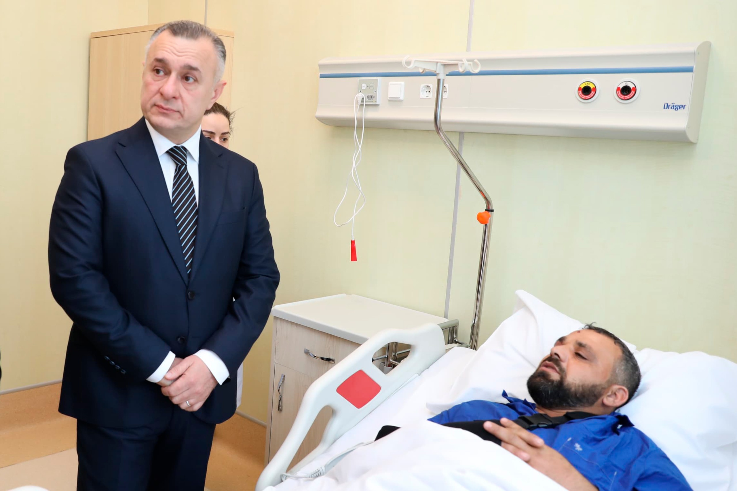 A man in a hospital bed looks unwell, while a suited individual stands nearby, appearing concerned.