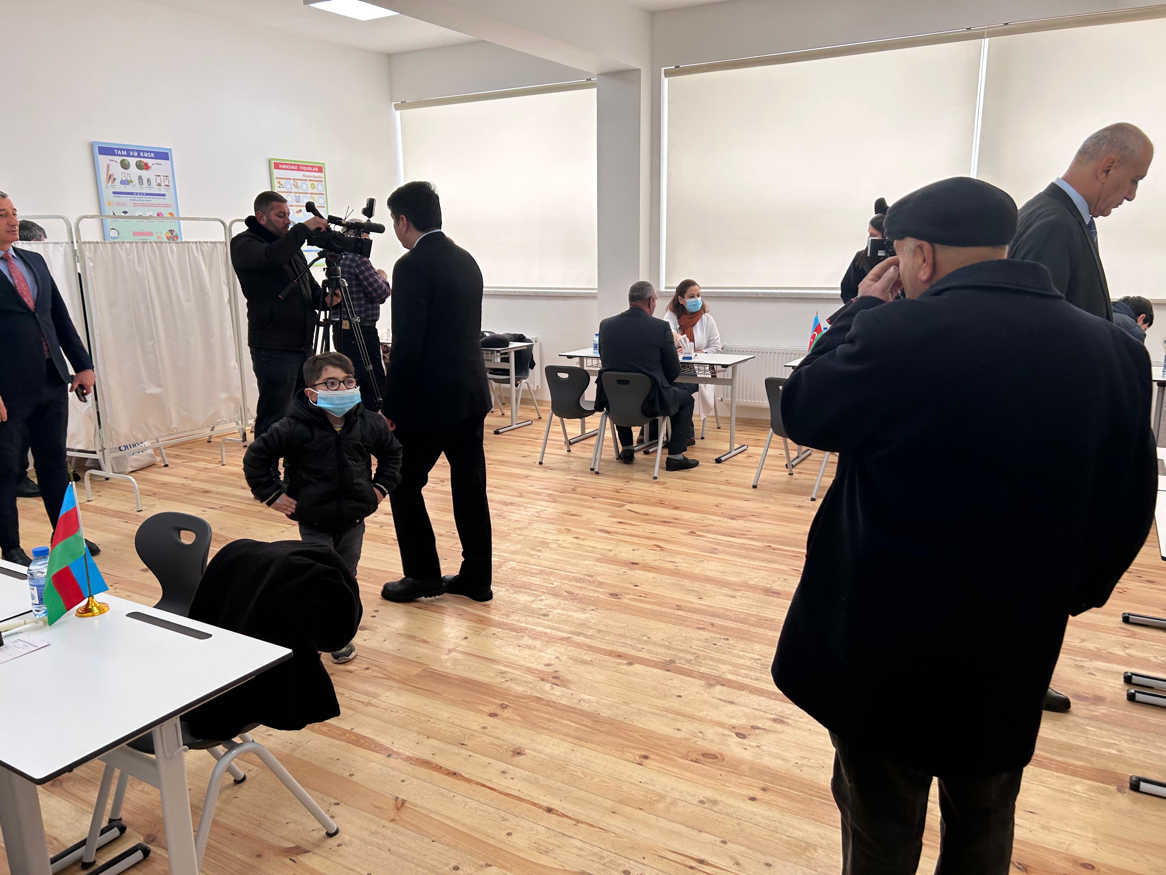 A classroom setting with people in suits, a child in a mask, and a camera crew. Flags and tables are present.