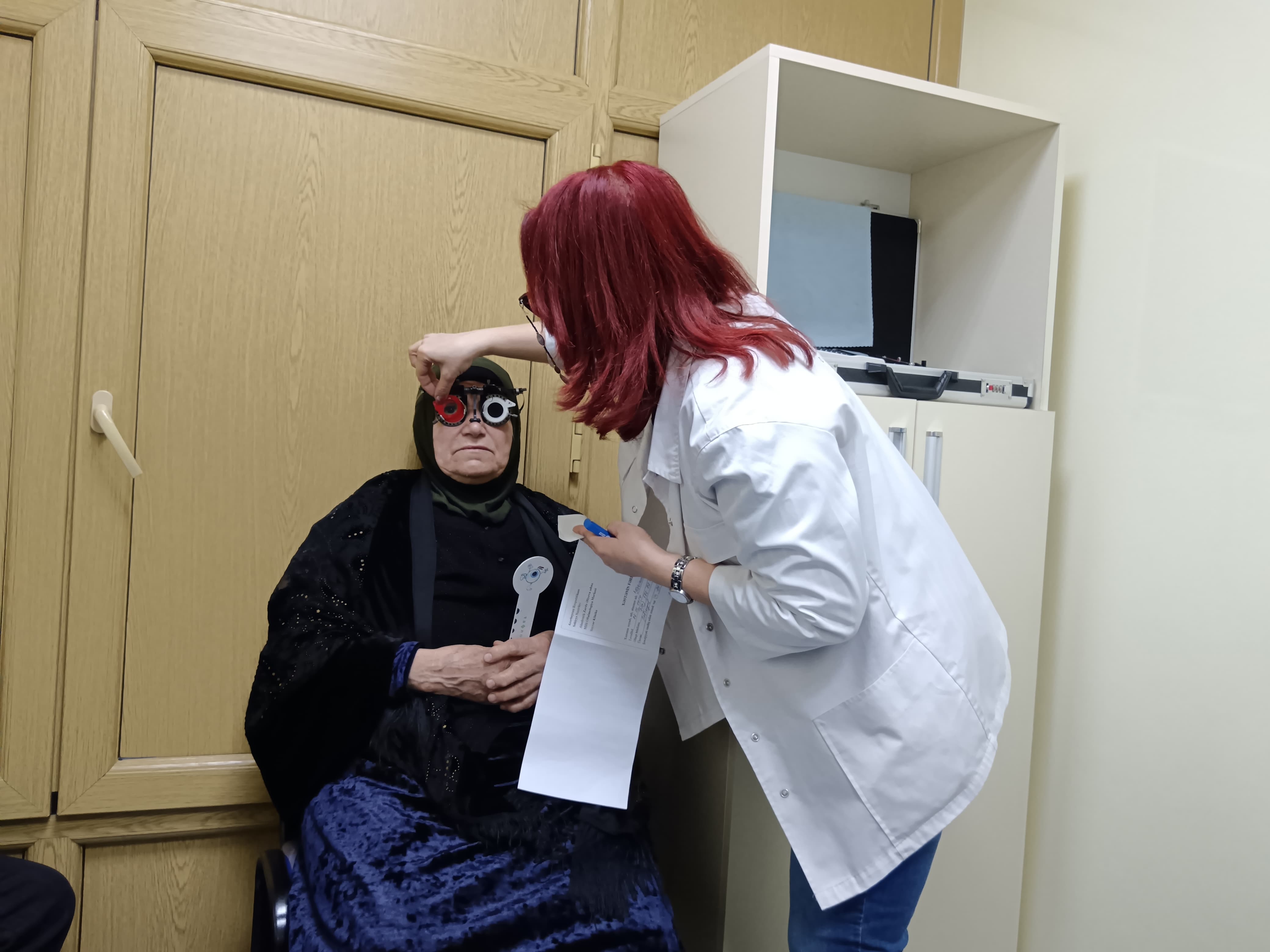 A woman in a black outfit sits while a healthcare professional adjusts vision testing glasses on her.