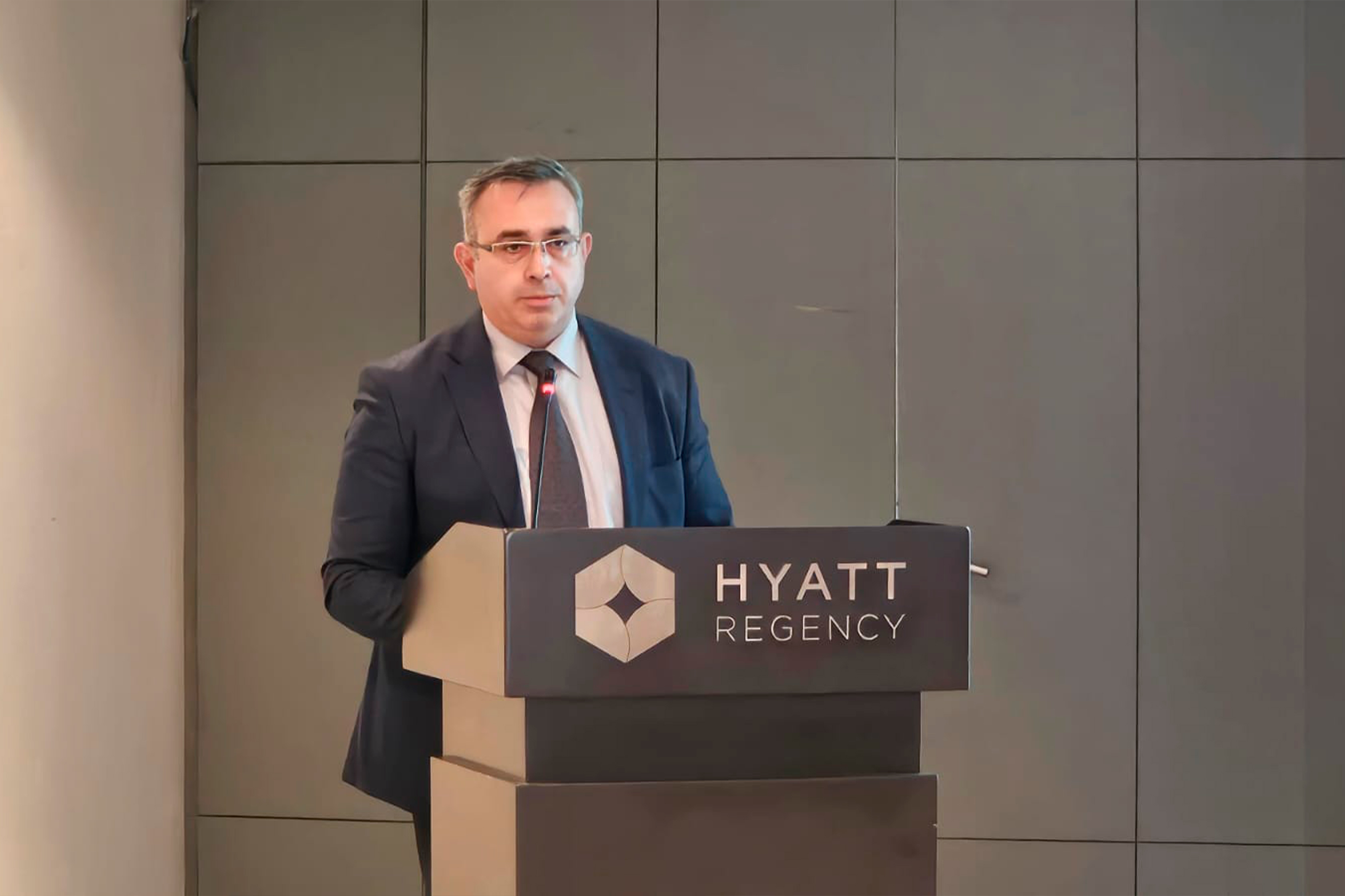 A man in a suit stands at a podium with a microphone, speaking at a Hyatt Regency event.