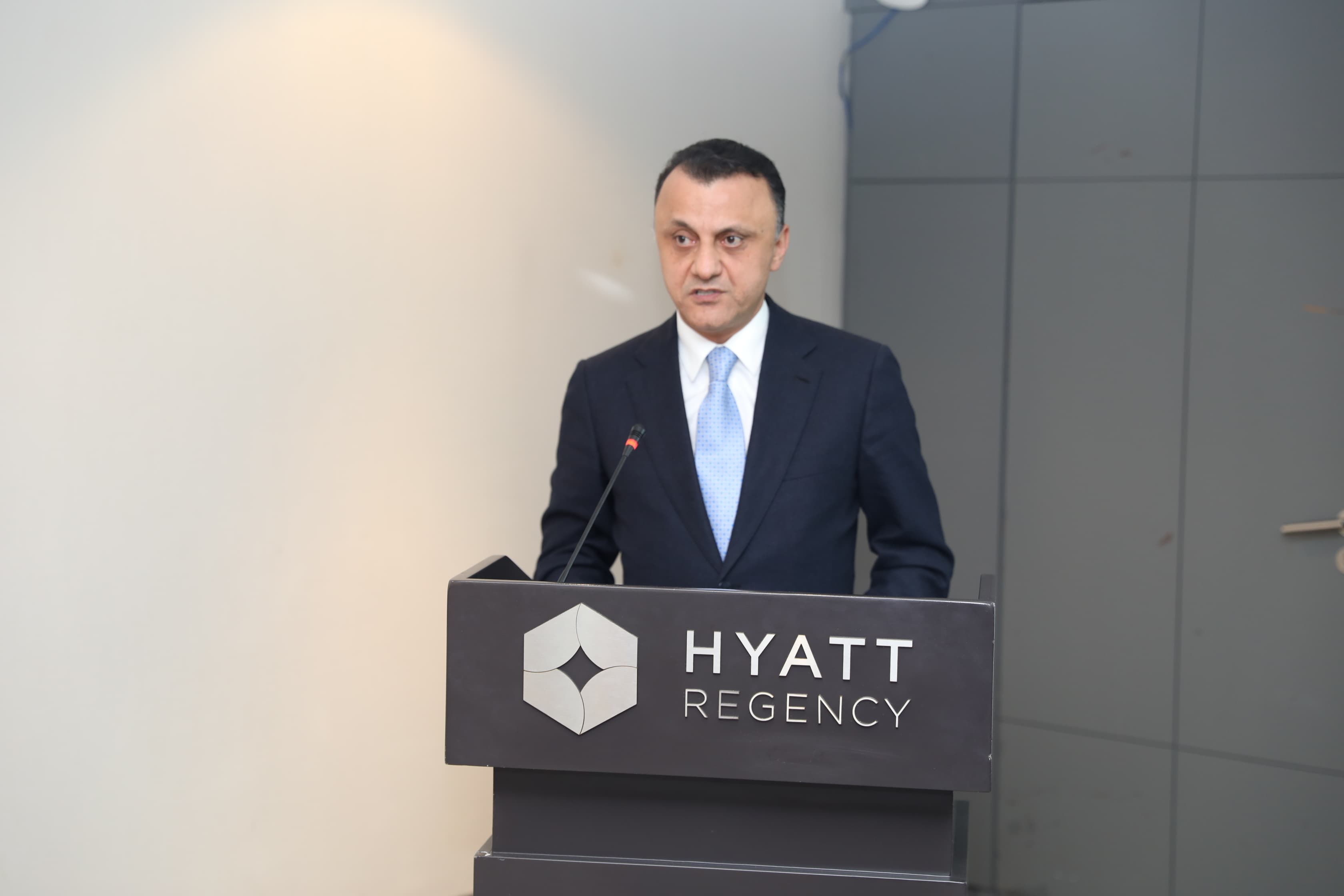 A man in a suit speaks at a podium with the Hyatt Regency logo, set against a neutral background.