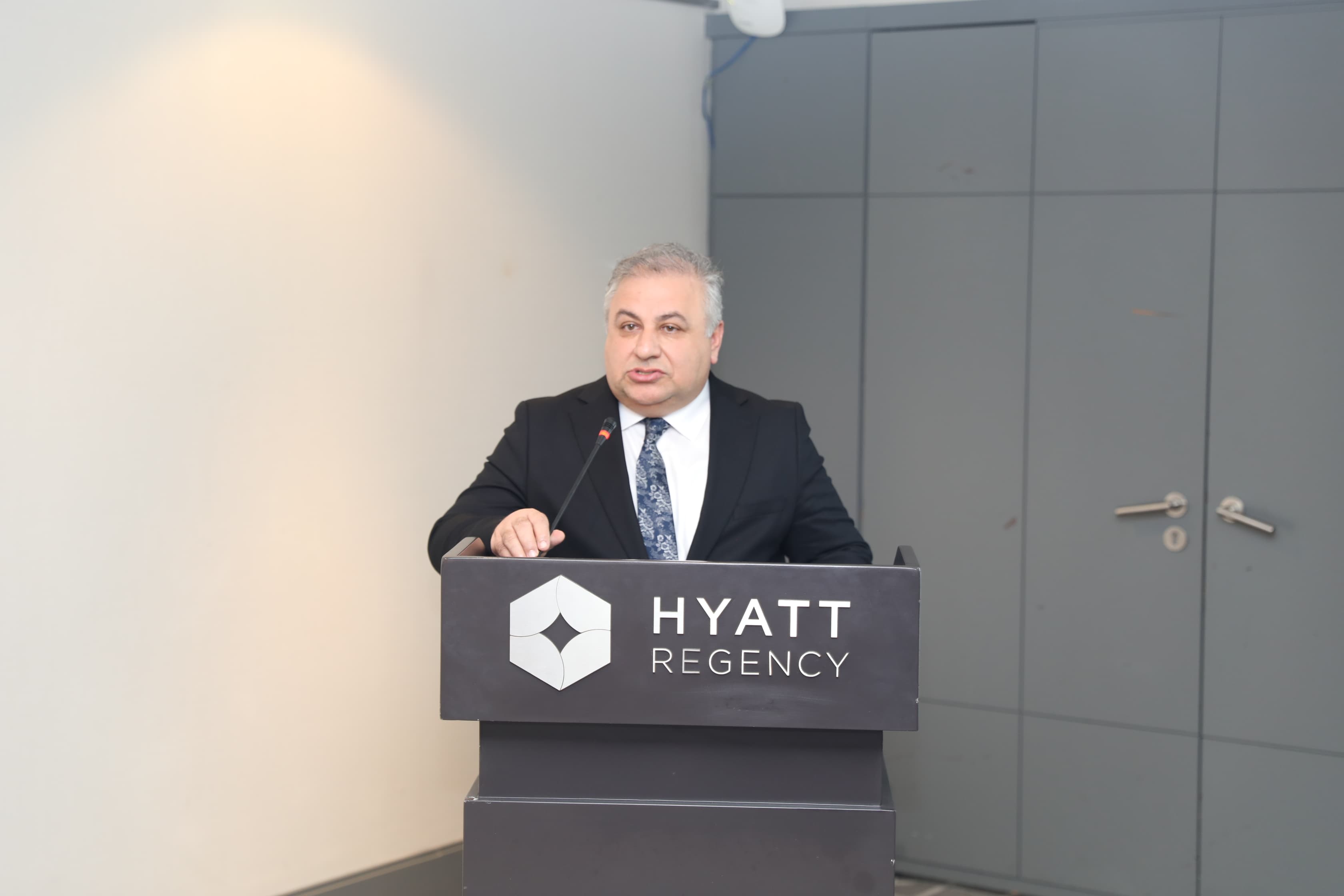 A man in a suit speaks at a podium with the Hyatt Regency logo, set against a neutral background.