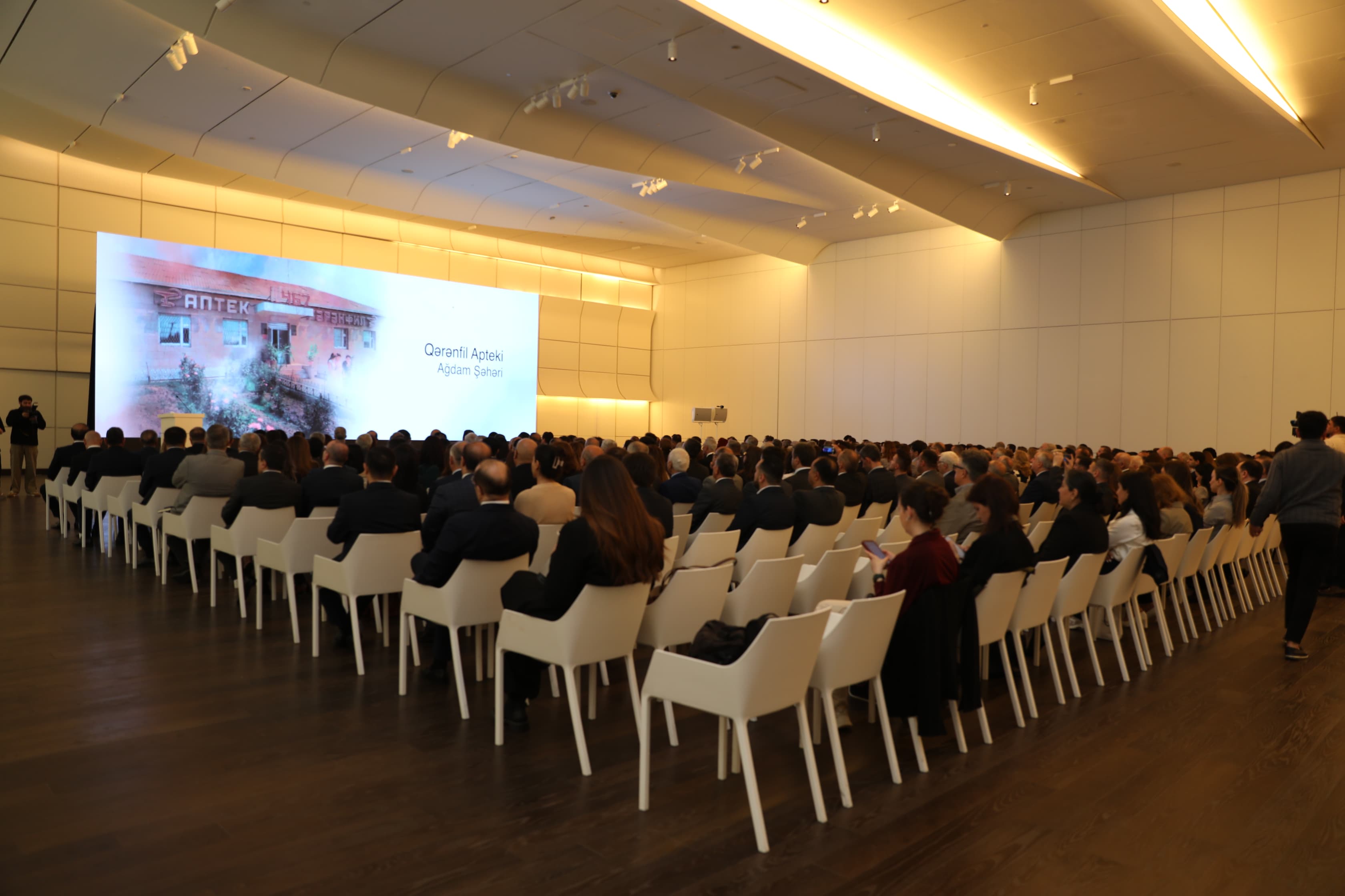 A large audience seated in a modern conference hall, facing a presentation screen displaying "Qərənfil Apteki, Ağdam Şəhəri."