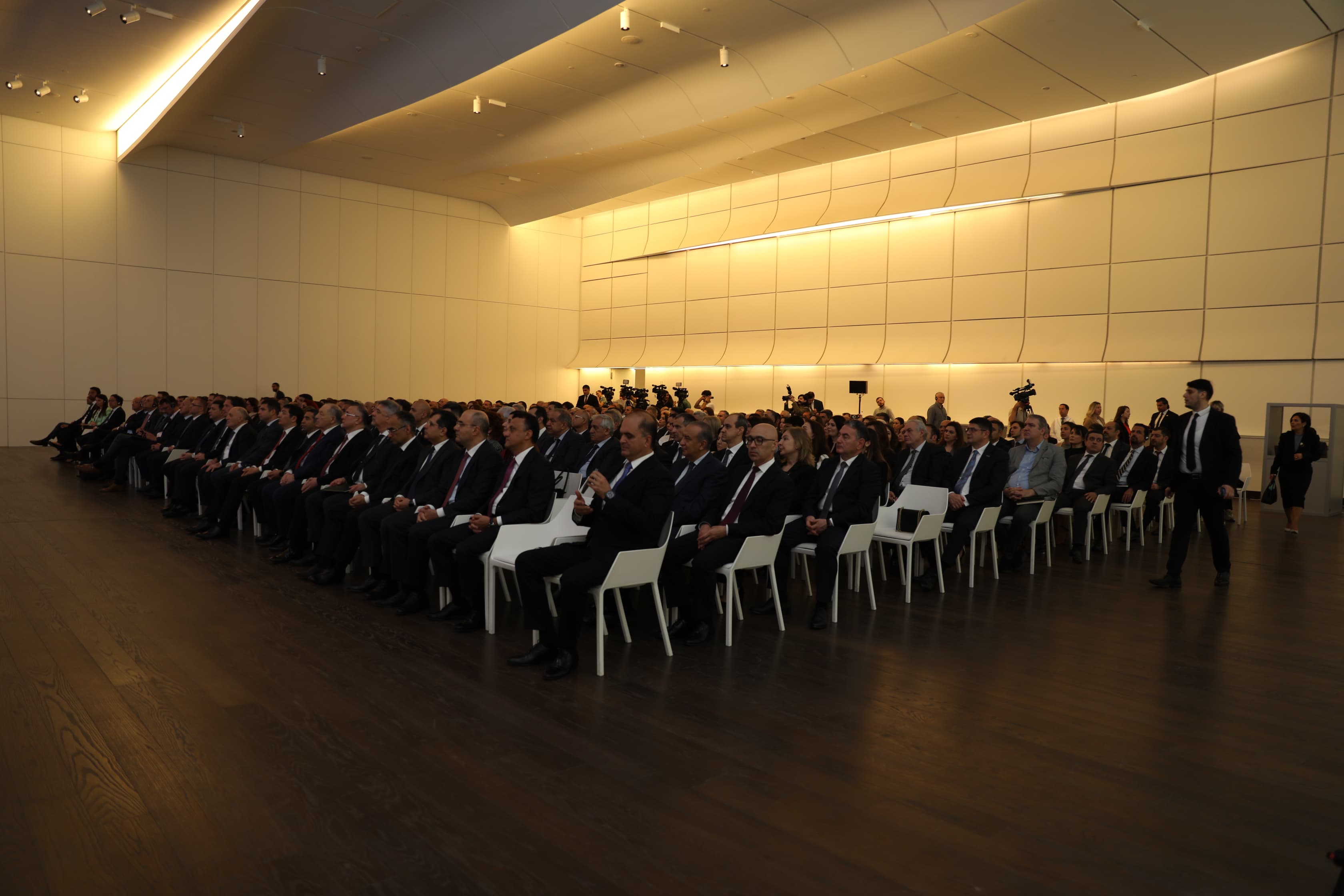 A large audience seated in a modern conference hall, attentively listening to a speaker.