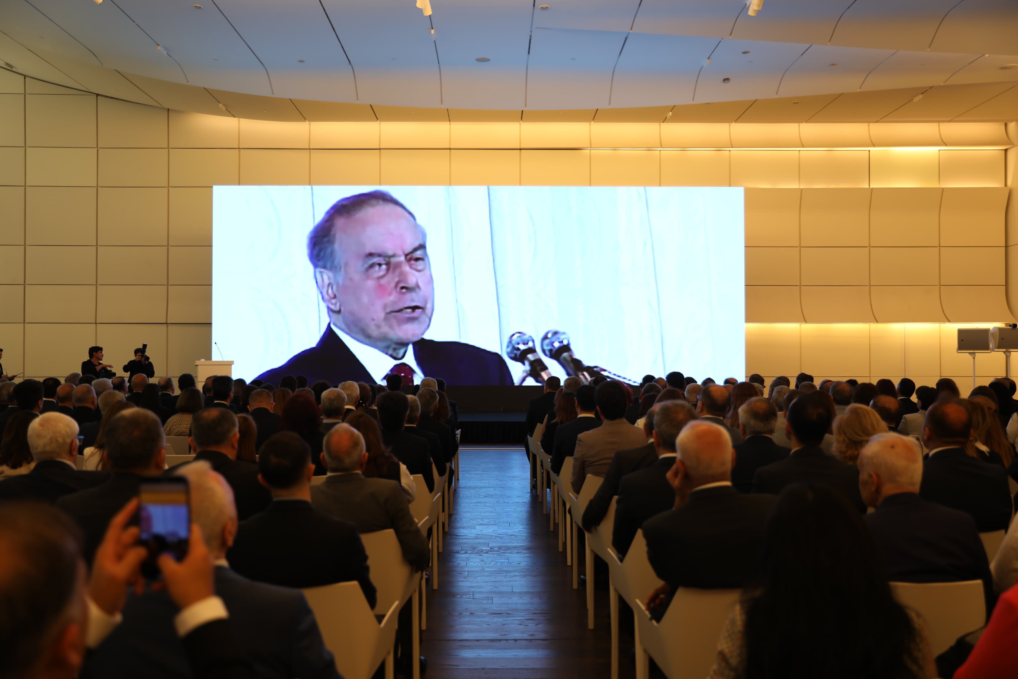 A large audience watches a speaker on a big screen at a formal event, with a modern venue and bright lighting.