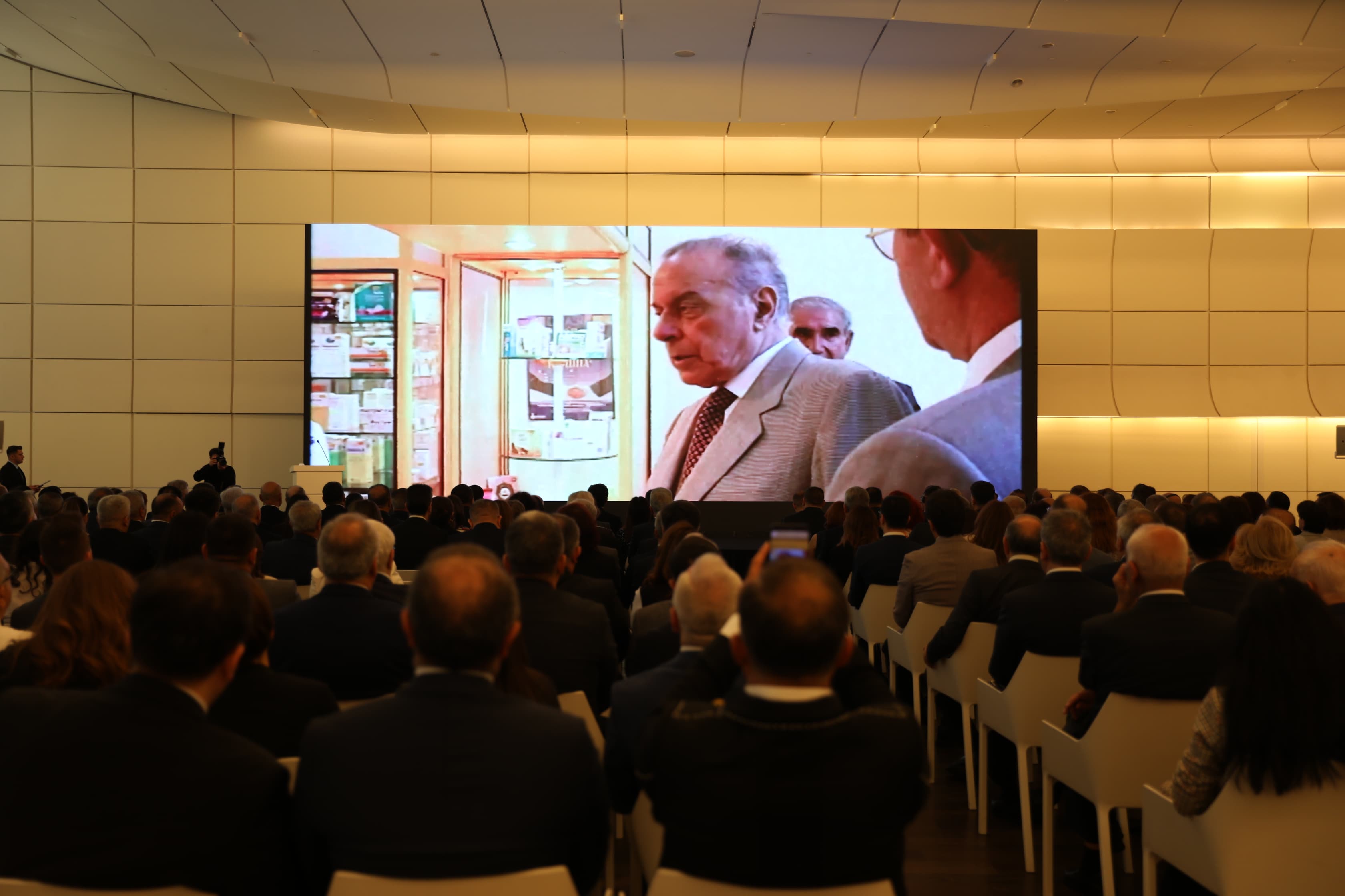 A large audience watches a presentation on a big screen, featuring a man in a suit and a display case behind him.