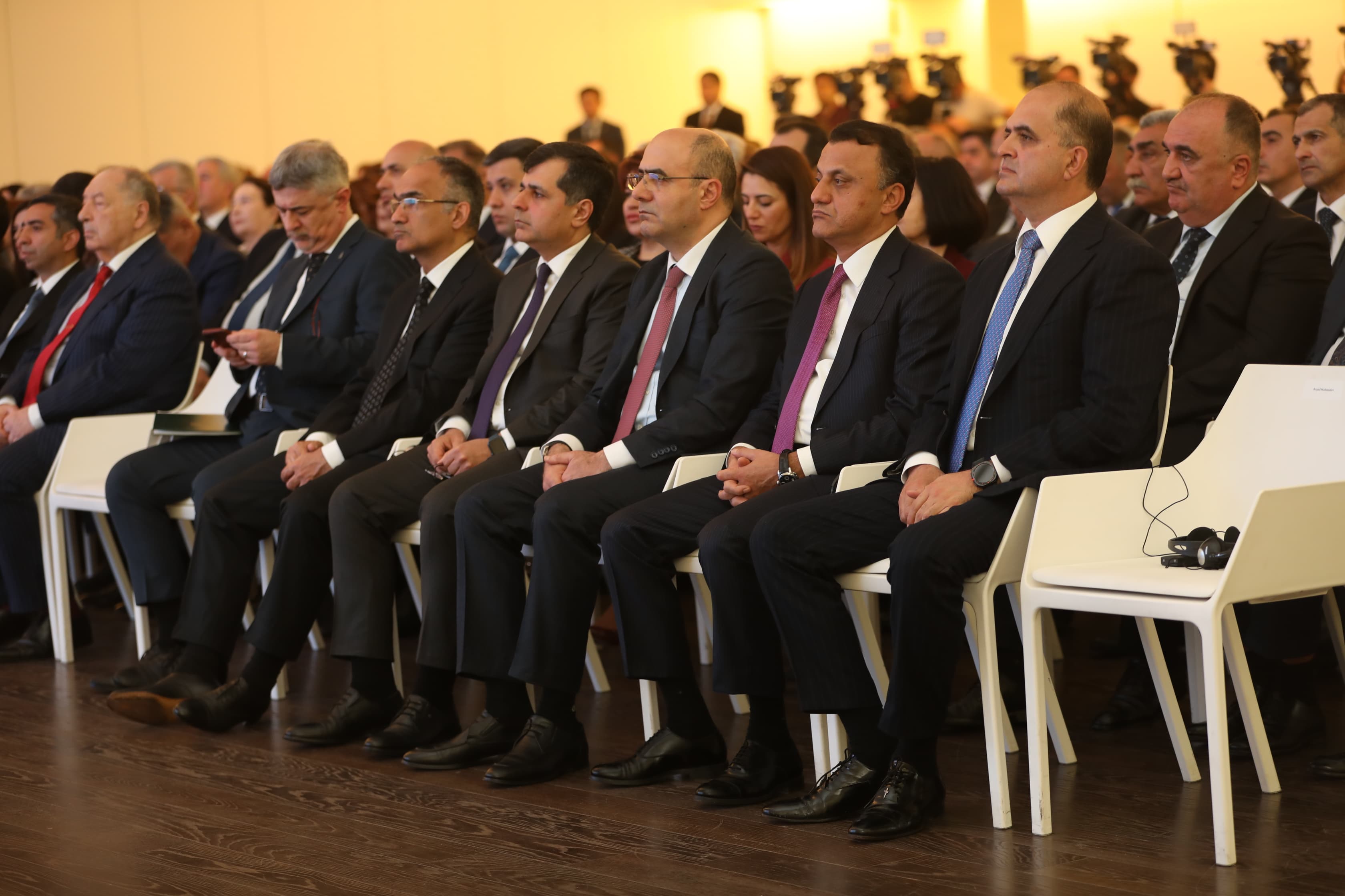 A group of men in formal attire seated in a row at an event, with a few women visible in the background.