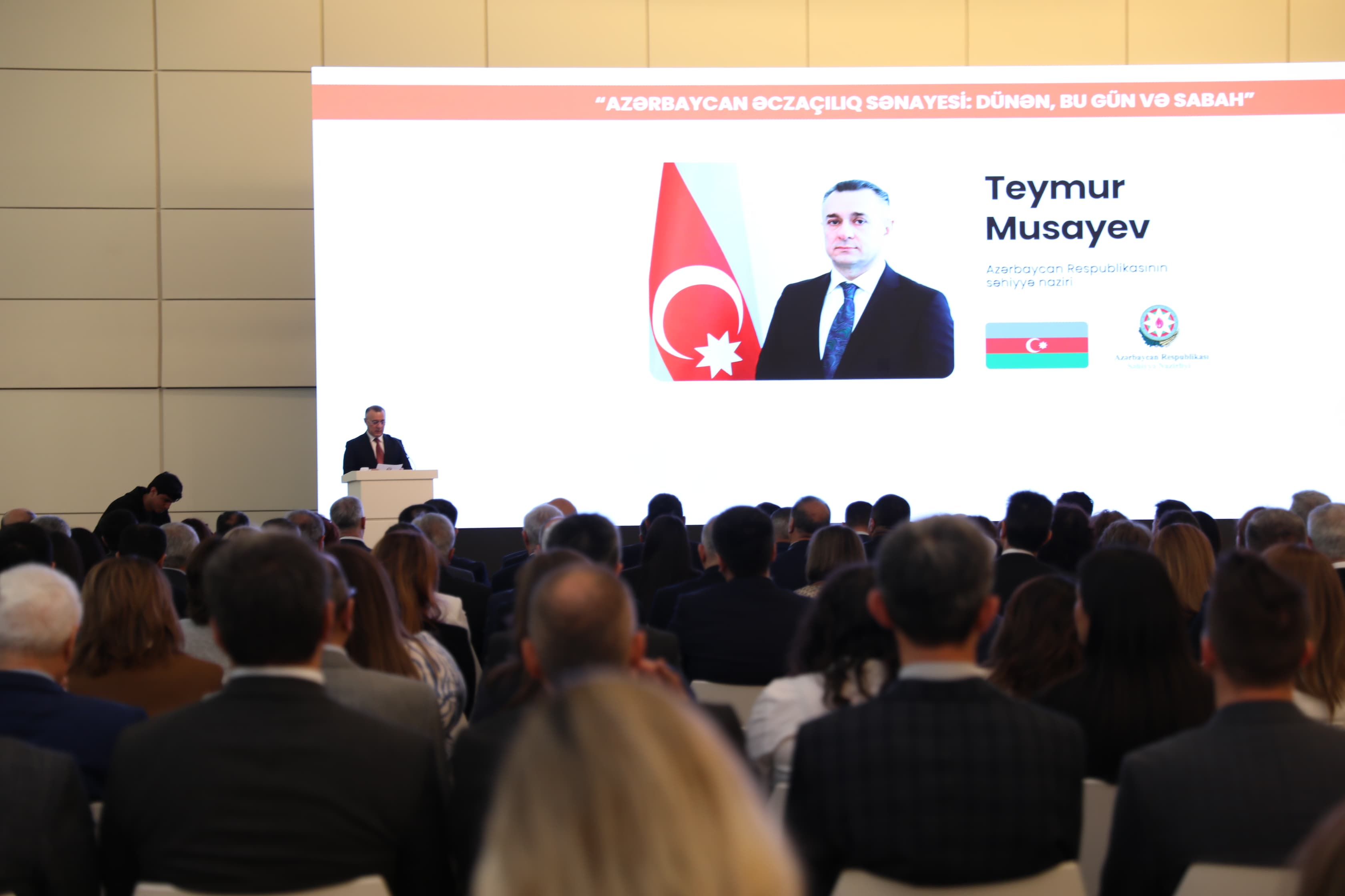A speaker presents at a conference, with an audience facing a large screen displaying information about Teymur Musayev.