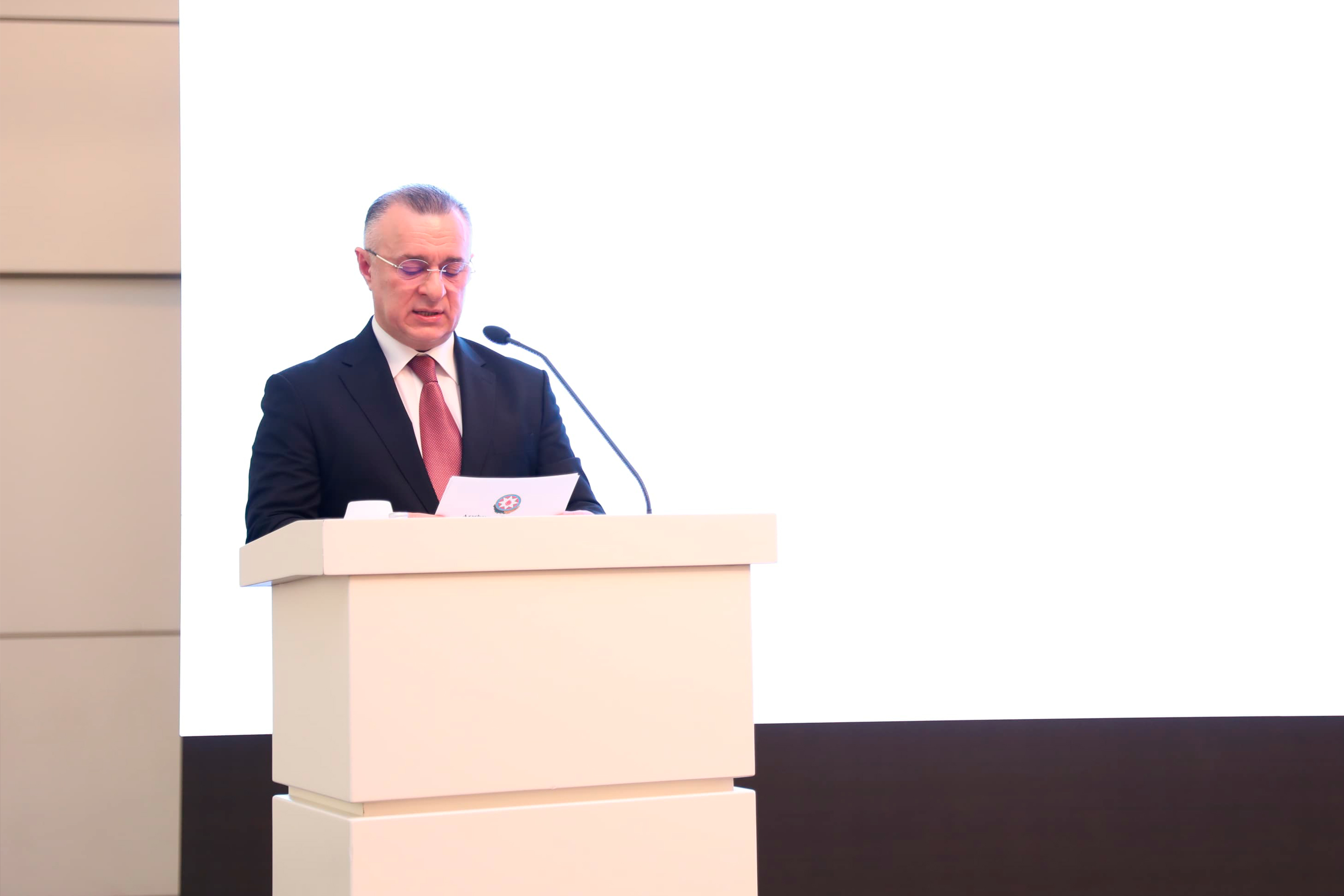 A man in a suit stands at a podium, reading from a paper, with a blank white background behind him.