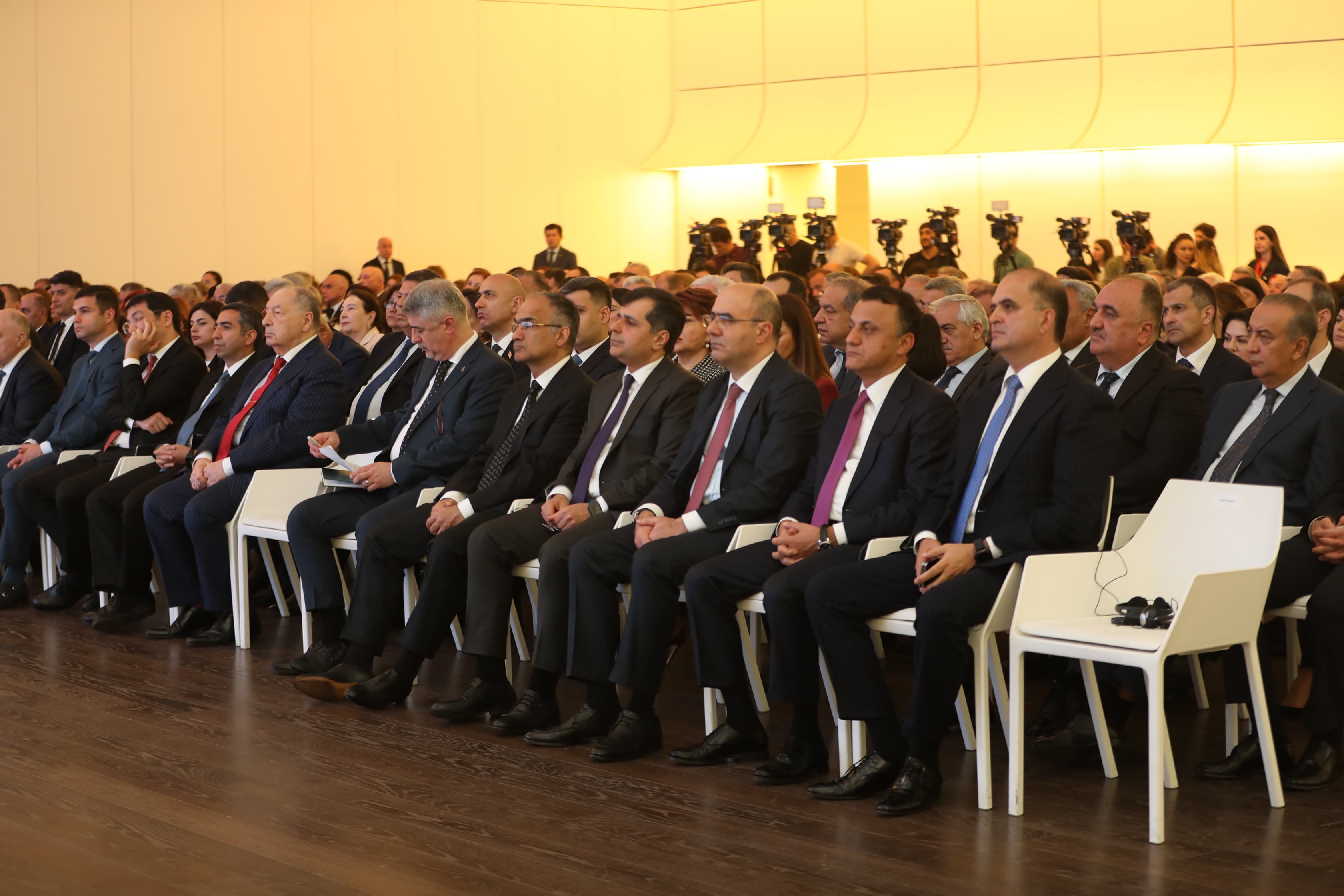 A large group of people in formal attire seated in rows, attending an event in a well-lit hall.