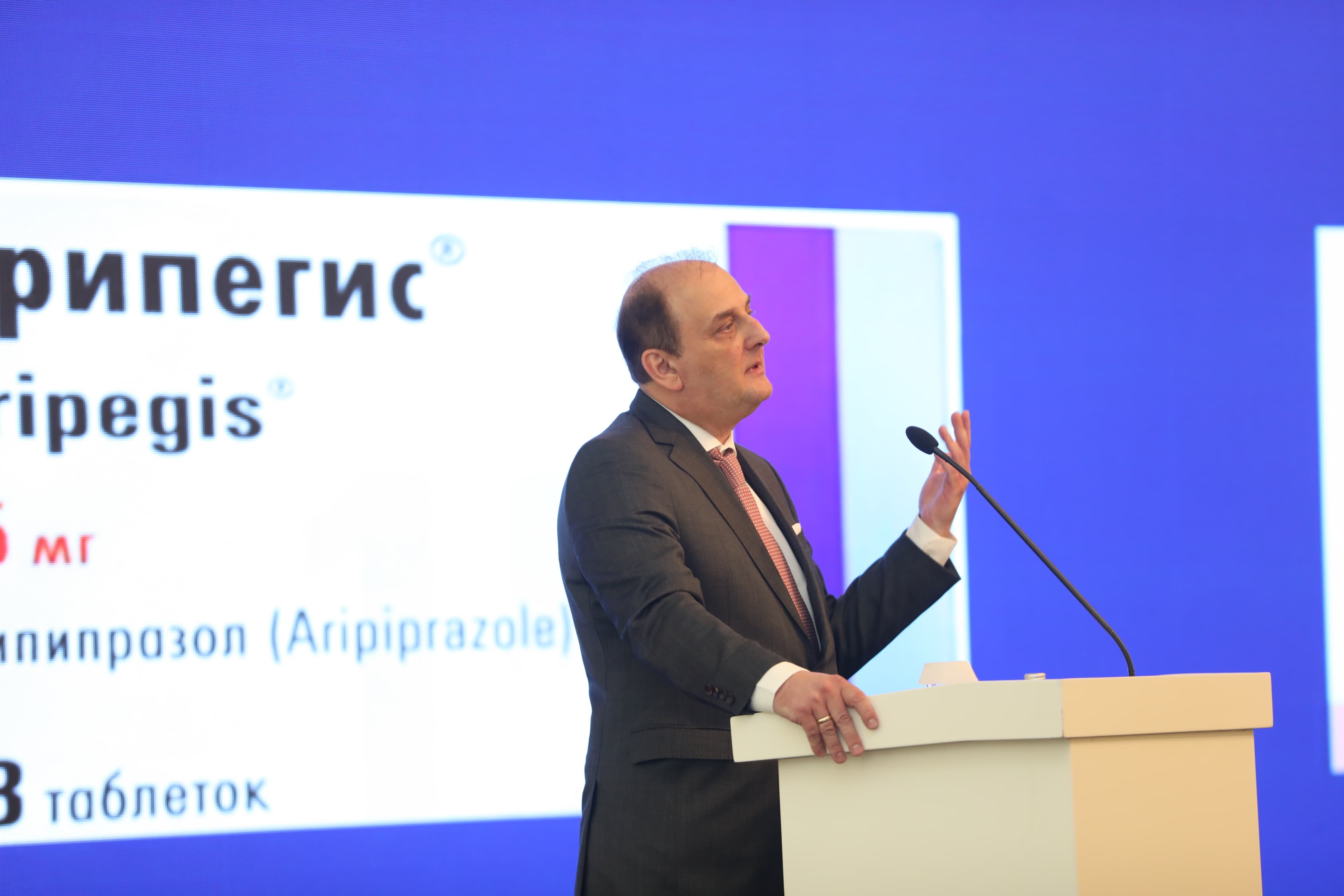 A speaker in a suit gestures while presenting at a podium, with a large screen displaying product information in the background.