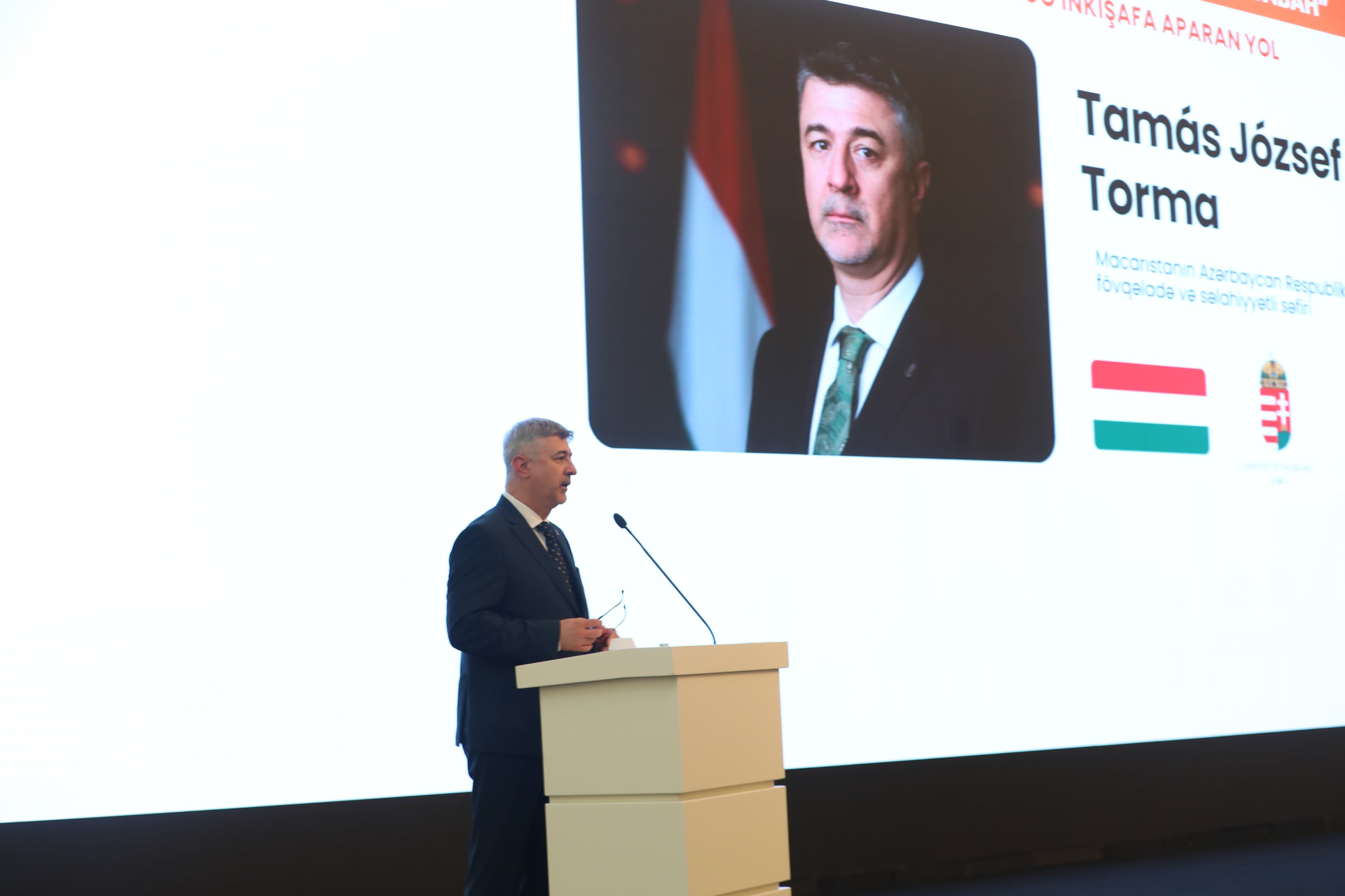 A man in a suit speaks at a podium, with a large screen displaying his photo and name in the background.