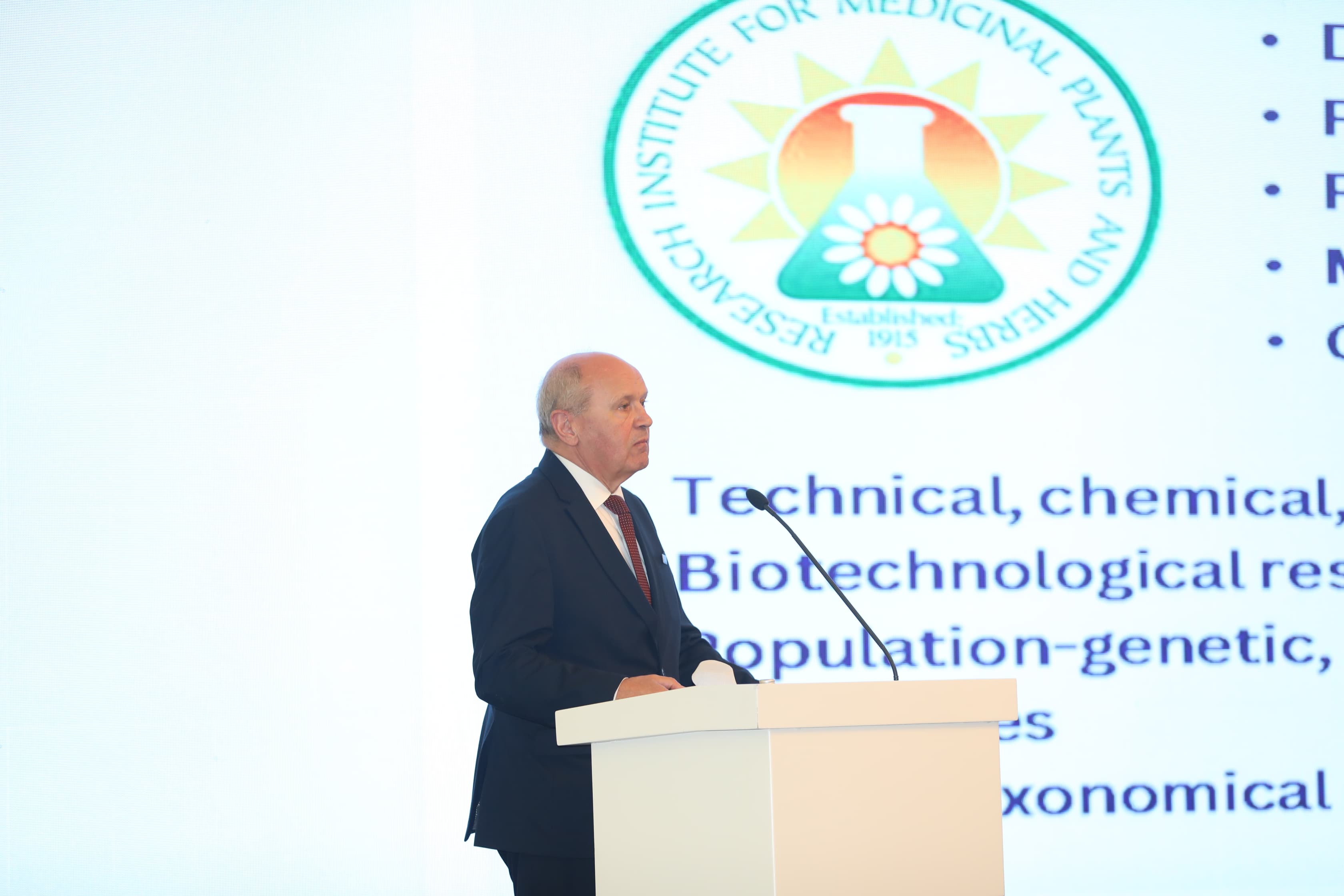 A speaker stands at a podium, presenting information about medicinal plants and herbs, with a logo in the background.