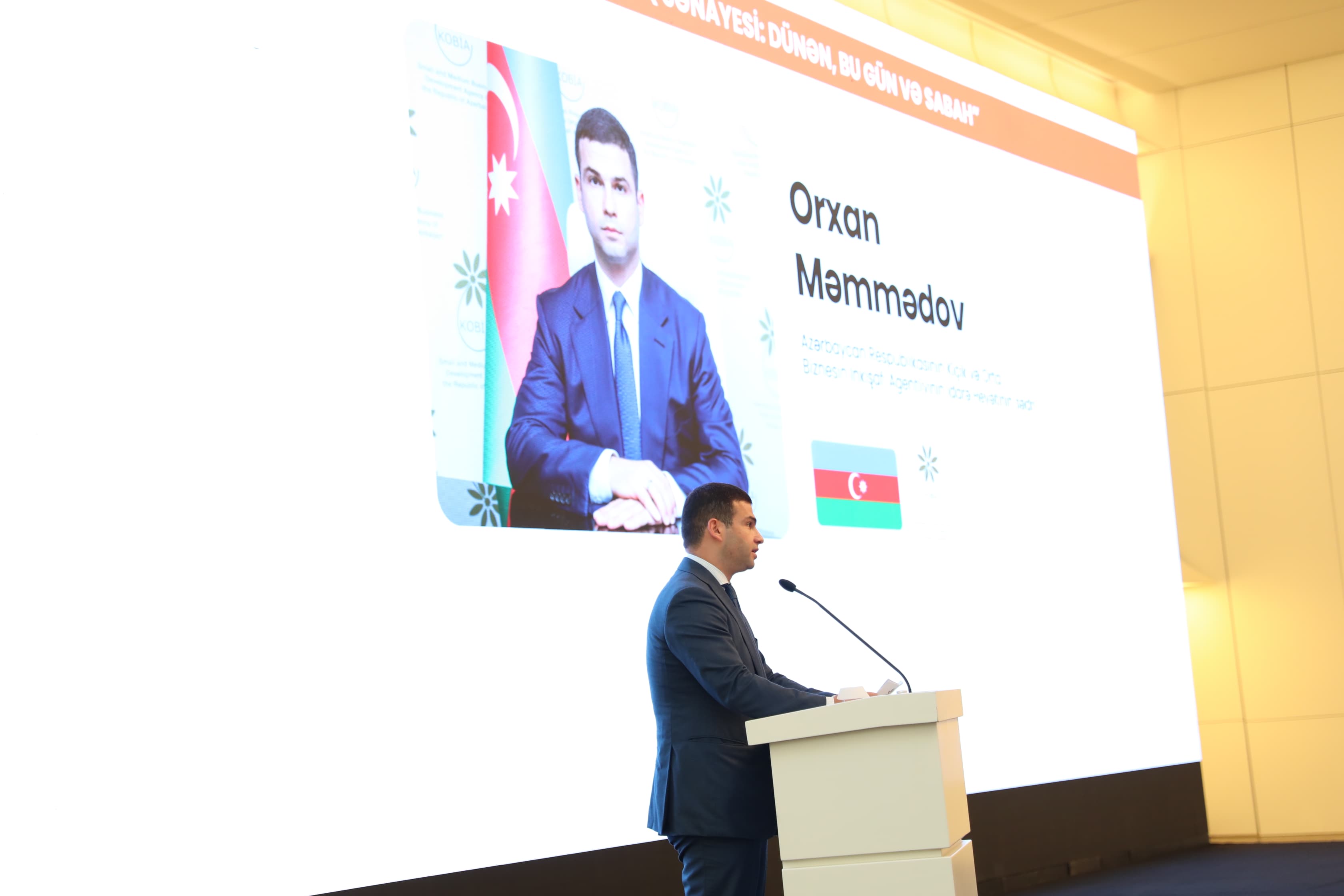A man in a suit speaks at a podium, with a large screen displaying his photo and information about him.