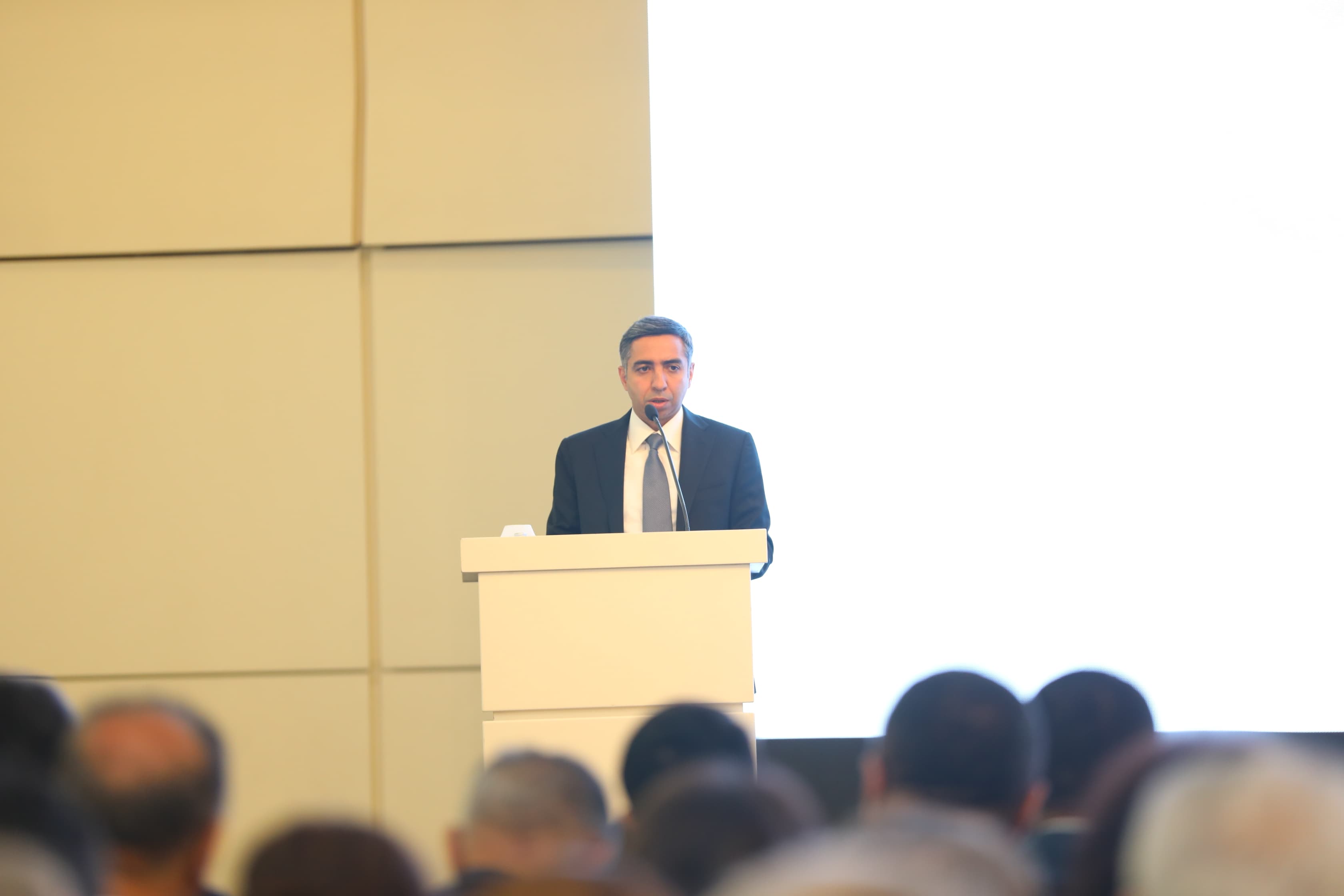 A speaker in a suit addresses an audience from a podium, with a blank screen behind him.