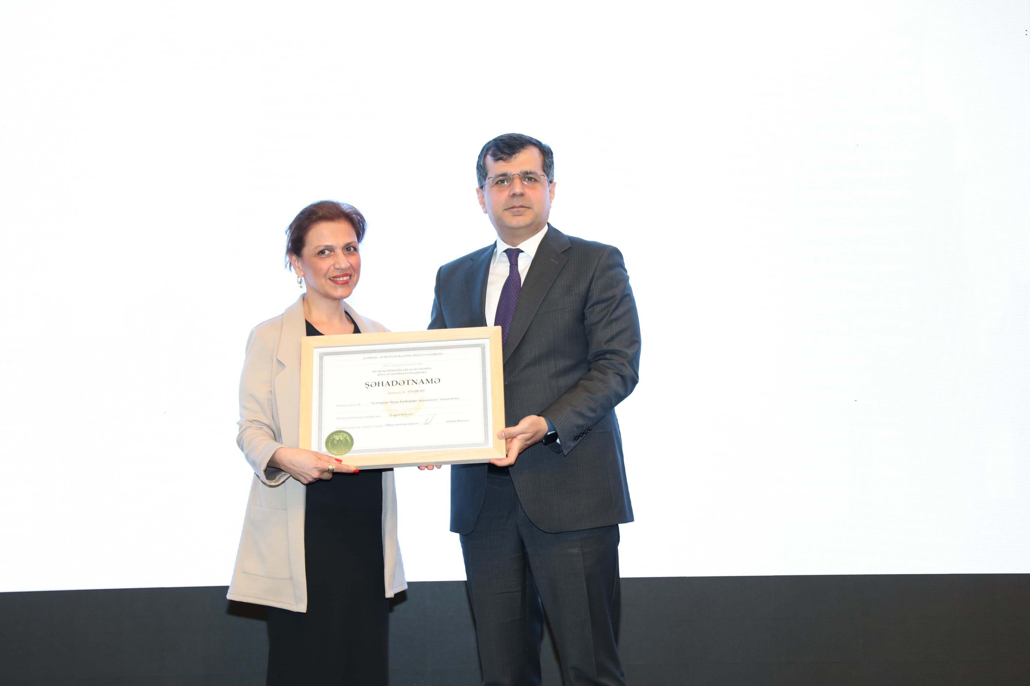 Two individuals are holding a certificate together, smiling. The background is mostly white, with a dark base.