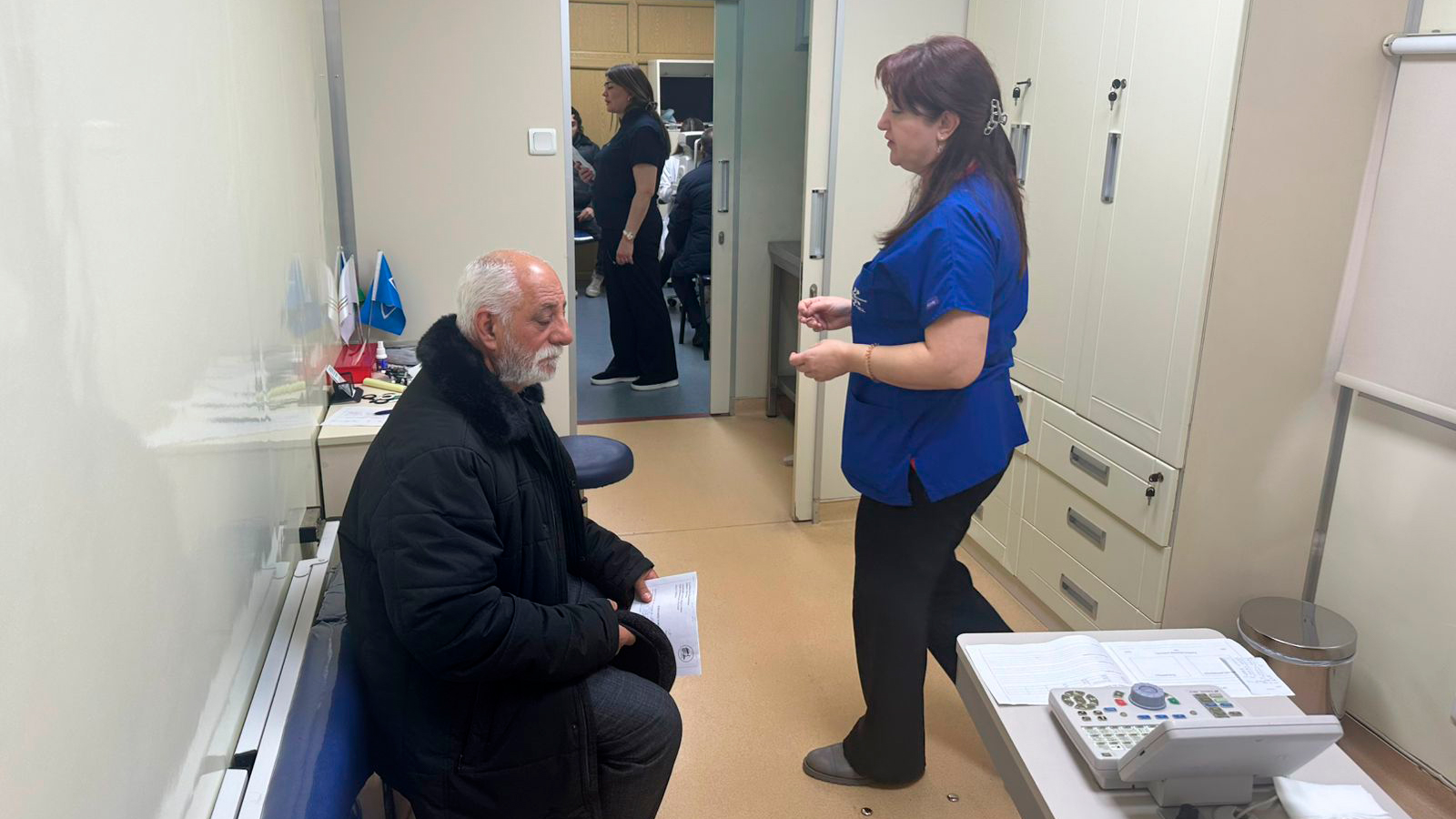 A man sits in a medical office while a woman in a blue shirt stands nearby, discussing something. Other staff are visible in the background.