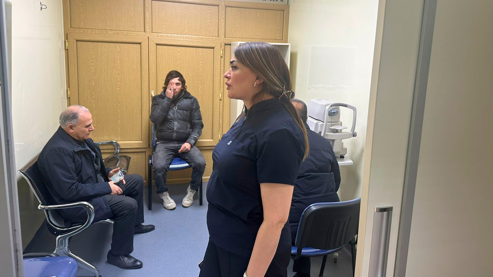 A woman stands in a medical office, while two men sit nearby. One man appears to be in thought, and medical equipment is visible.