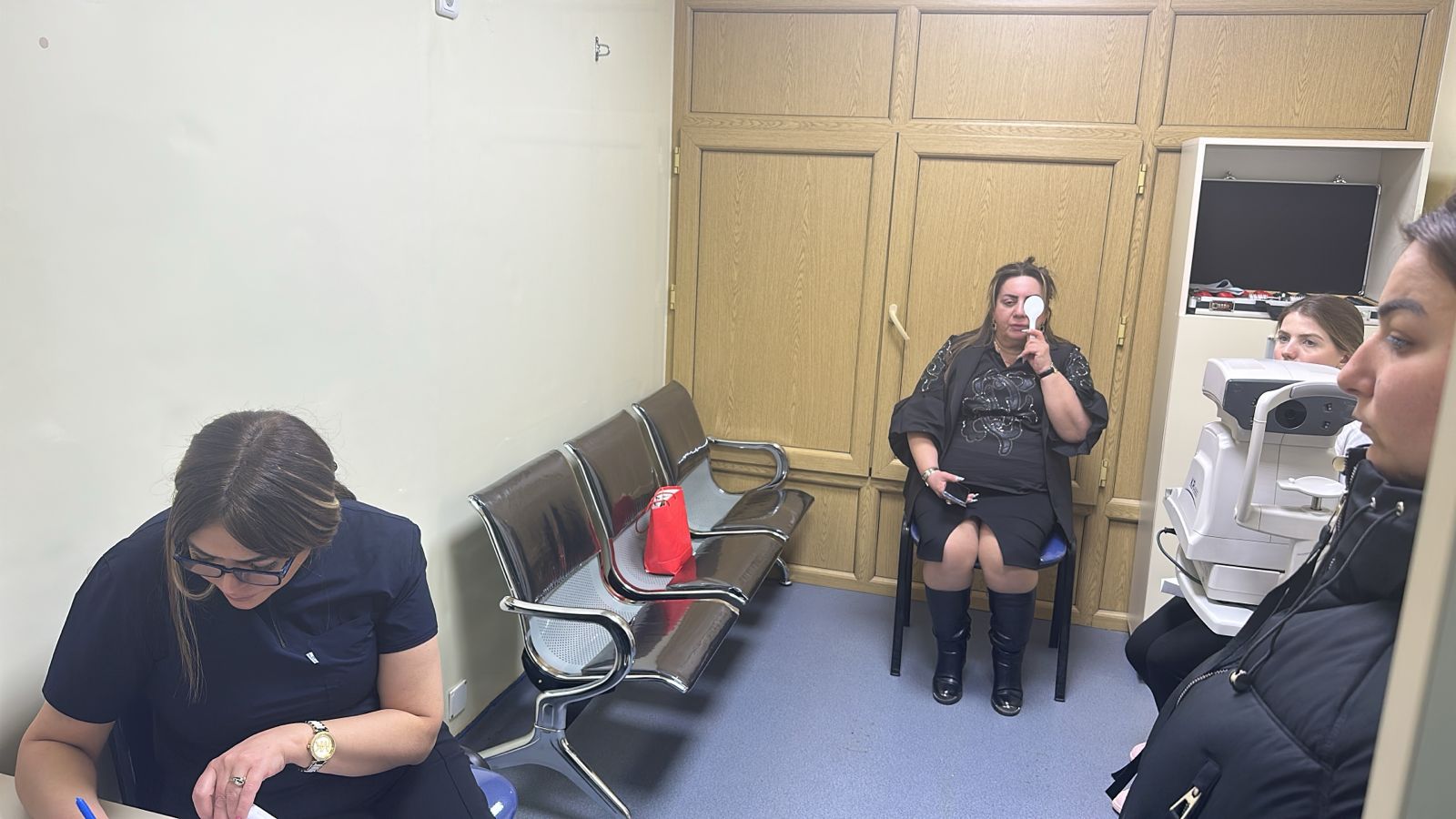 A medical office scene with four women. One is writing, while another is undergoing an eye exam. Chairs and medical equipment are visible.