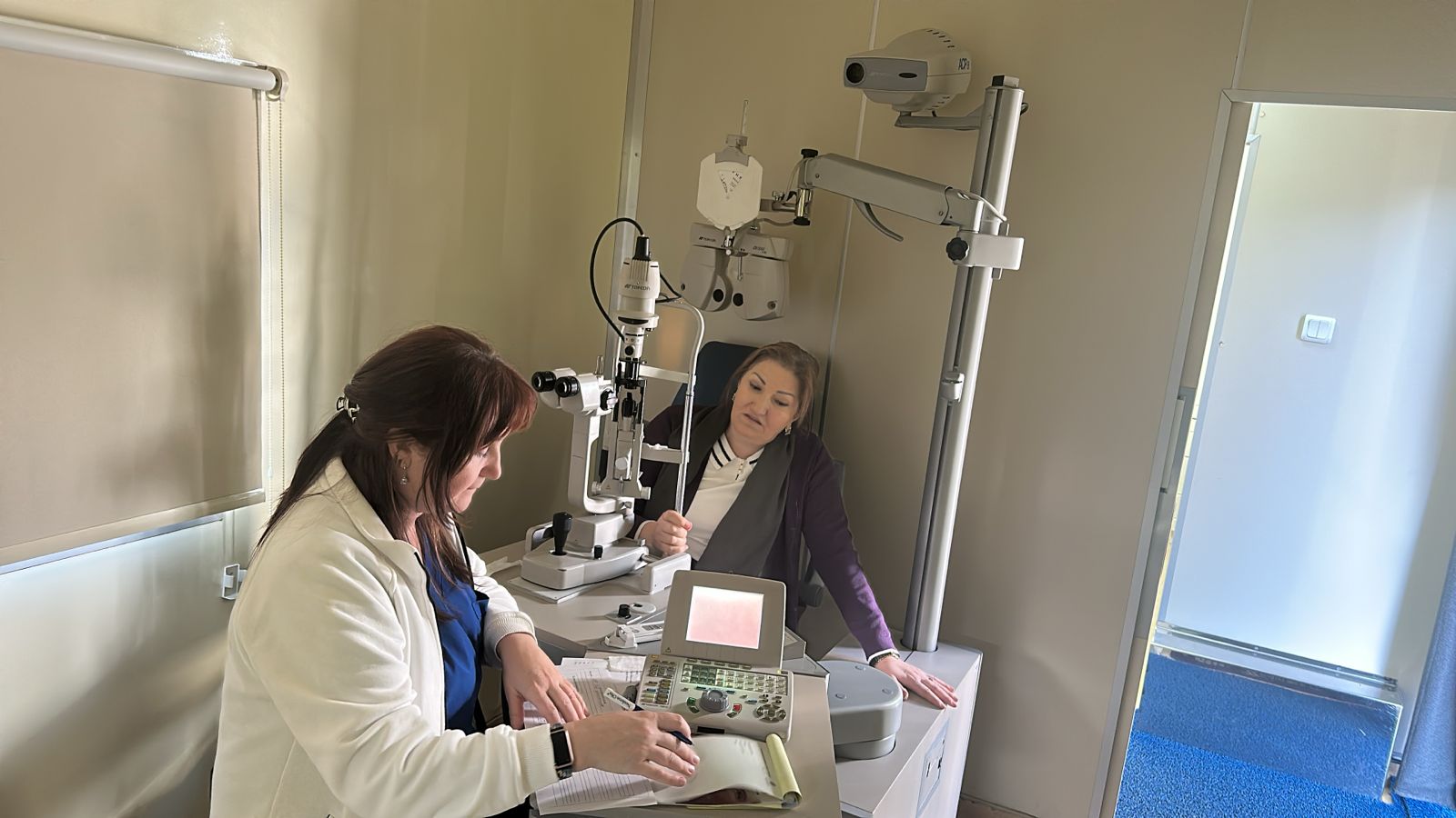 Two women are in a medical office, one using an eye examination machine while the other observes. Medical equipment is visible.